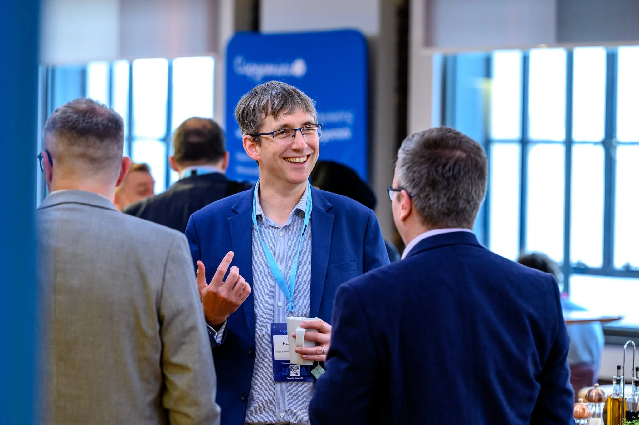 Three men engaged in conversation at a networking event, with the man in the center smiling and holding a coffee cup, wearing a blue blazer and glasses, in a bright room with large windows and other people in the background.