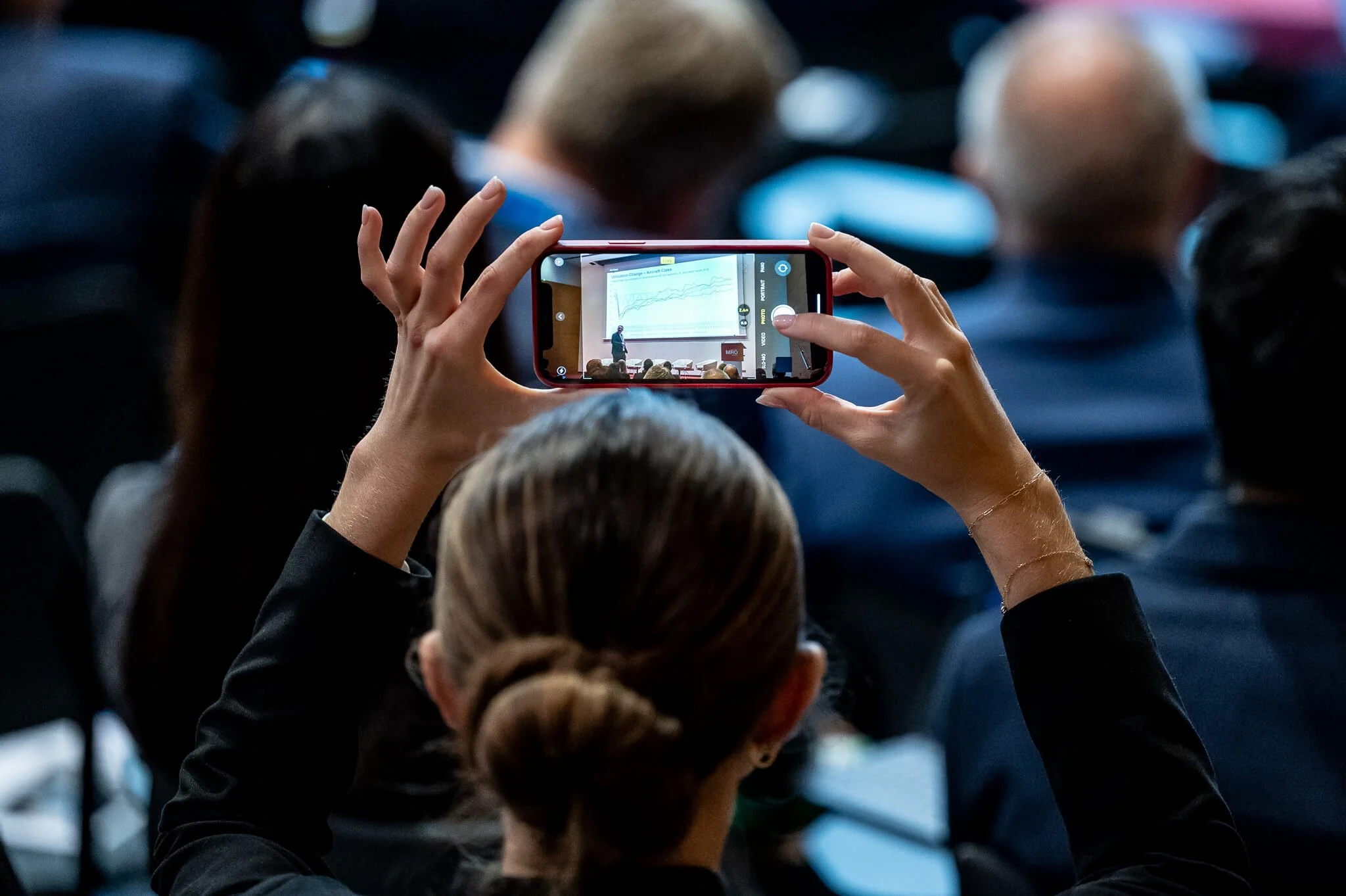 A woman with a bun is using her smartphone to record or take a picture of a presentation in a conference room, with a large screen displaying a graph in front of an audience.