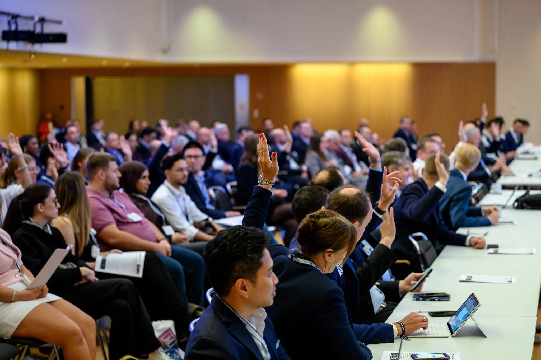 A large group of people attending a conference or meeting, with several individuals raising their hands.