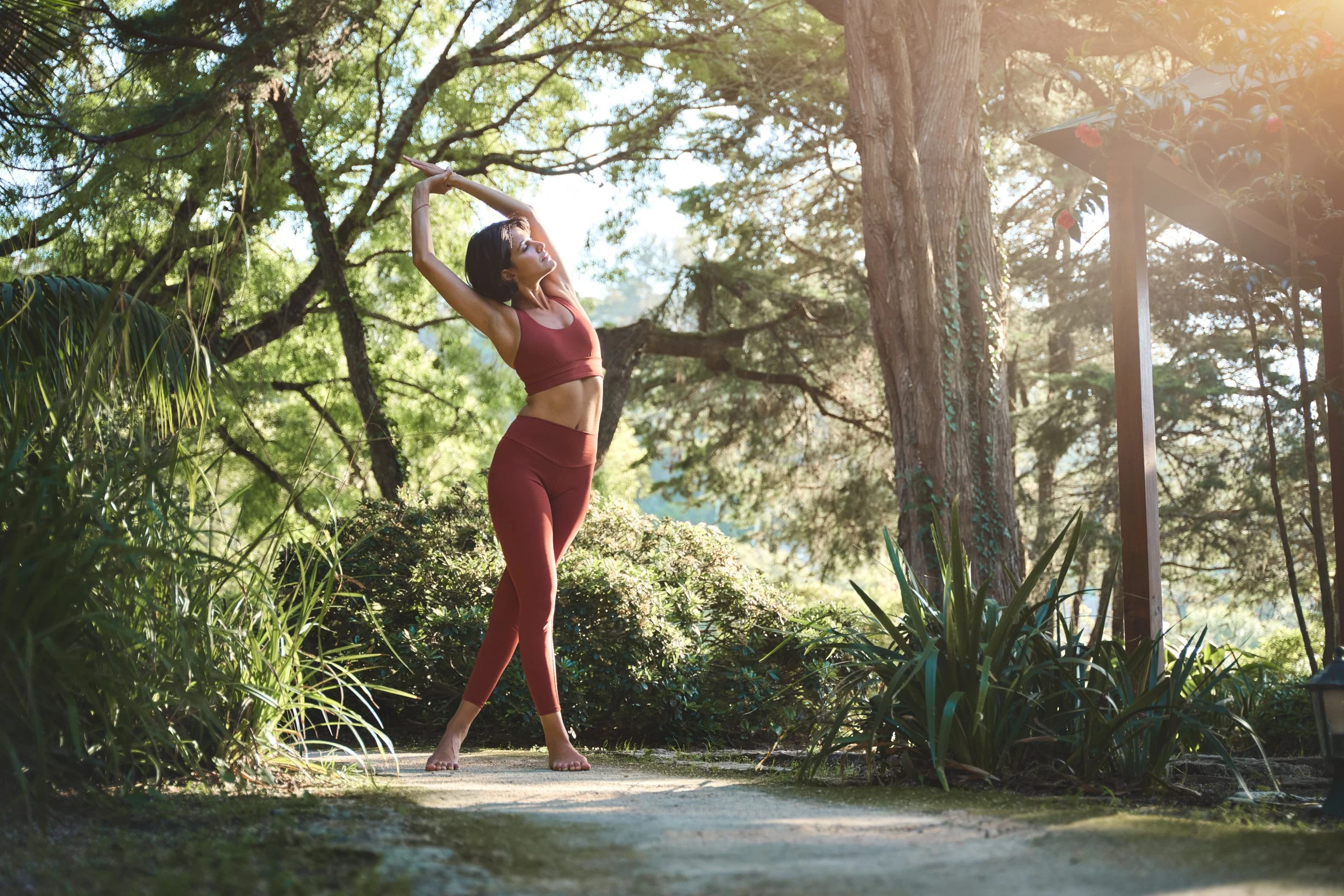 A woman in workout clothes stretching outdoors in a forested area with sunlight filtering through trees.