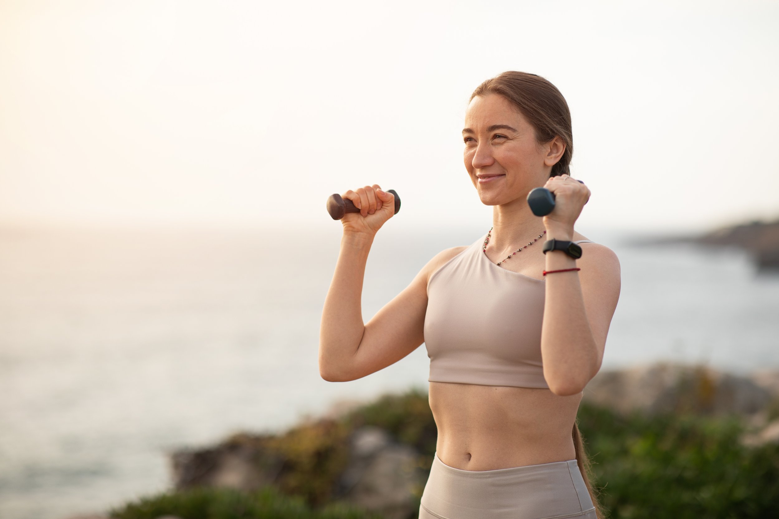 Woman exercising outdoors near the beach, holding dumbbells in both hands, smiling, during sunset or sunrise.