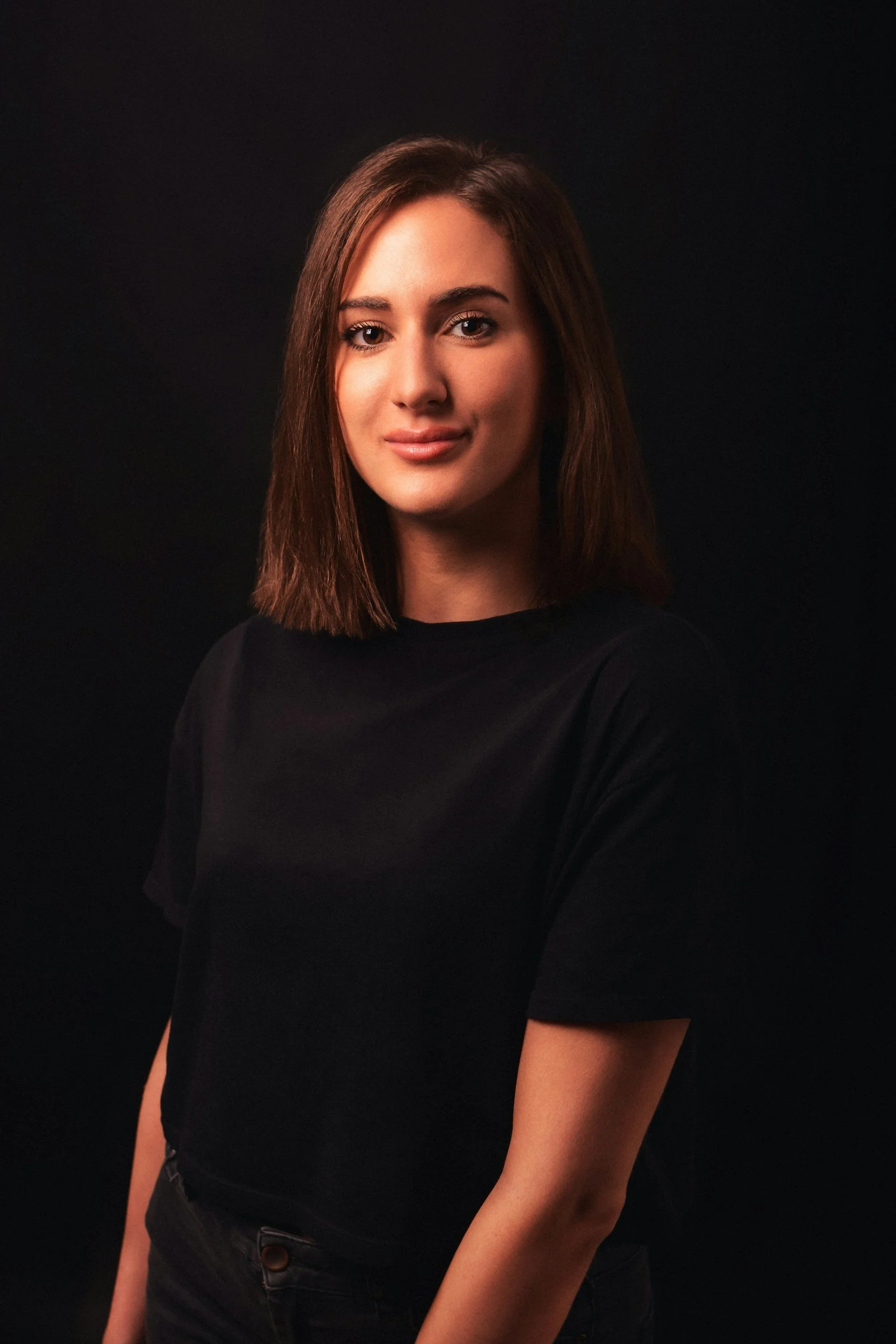 Portrait of a young woman with shoulder-length brown hair, wearing a black T-shirt, against a dark background.