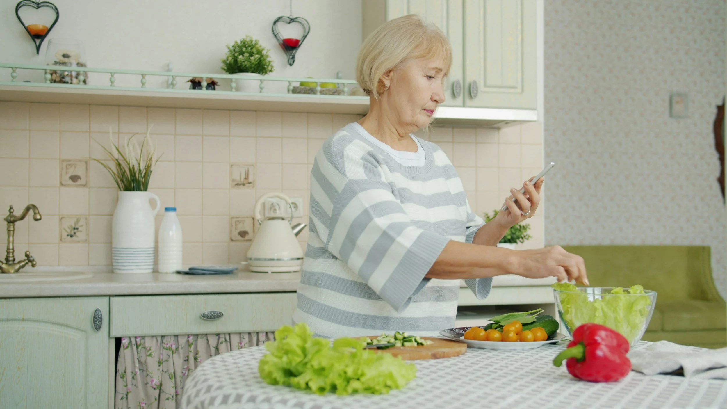 An elderly woman preparing a salad in a kitchen, surrounded by fresh vegetables including red bell peppers, cherry tomatoes, cucumbers, and lettuce, with kitchen cabinets, a sink, and decorative wall hangings in the background.