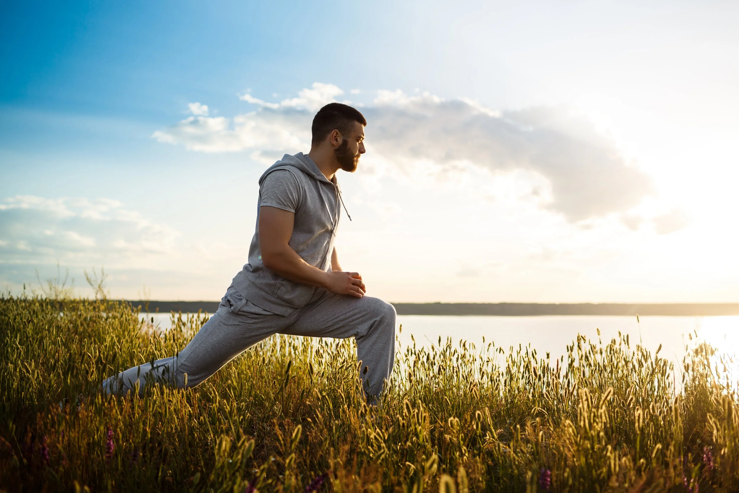 Man doing yoga in a grassy field near a body of water during sunset.