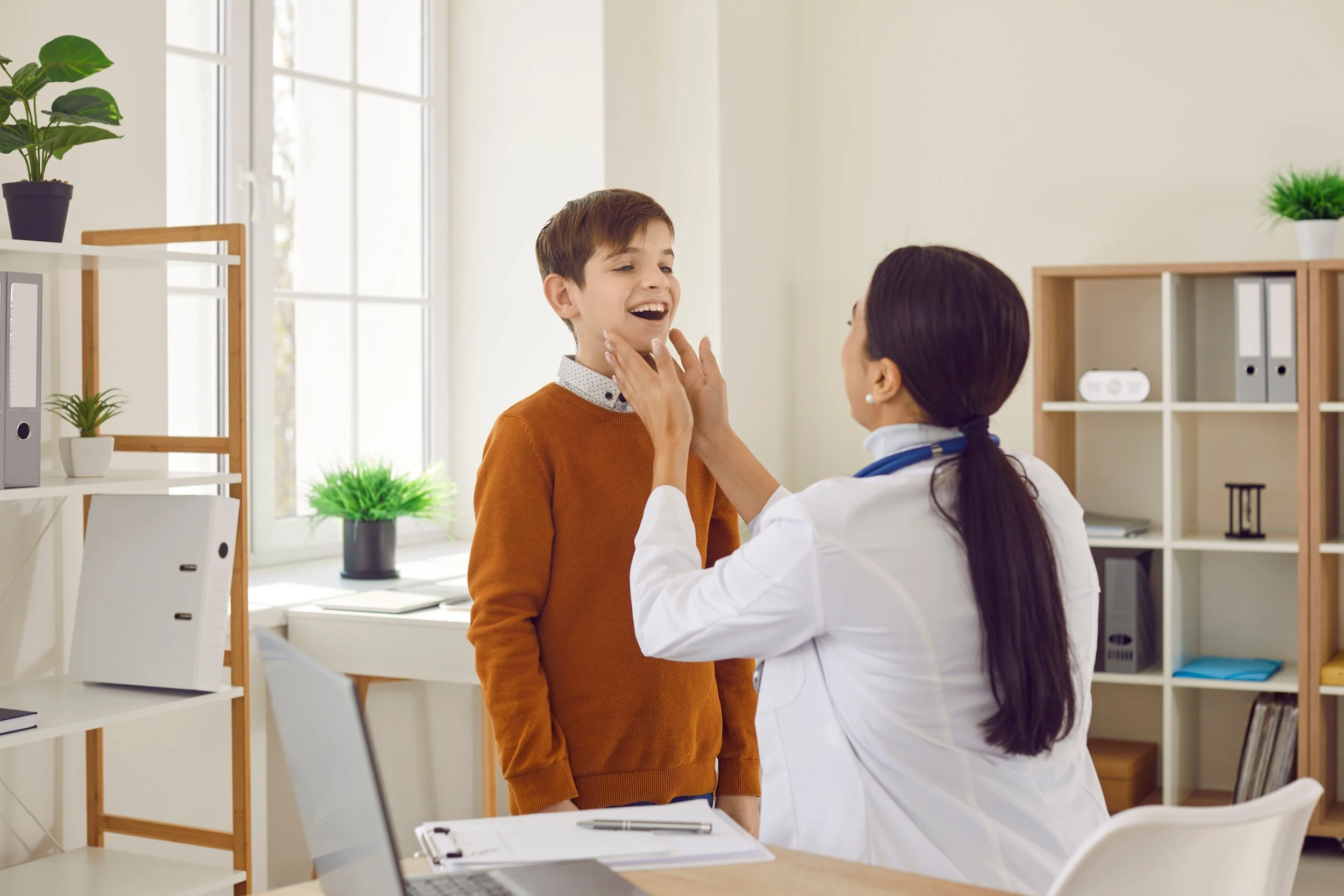 A female doctor examines a young boy in a medical office, using her hands on his chin while the boy smiles.
