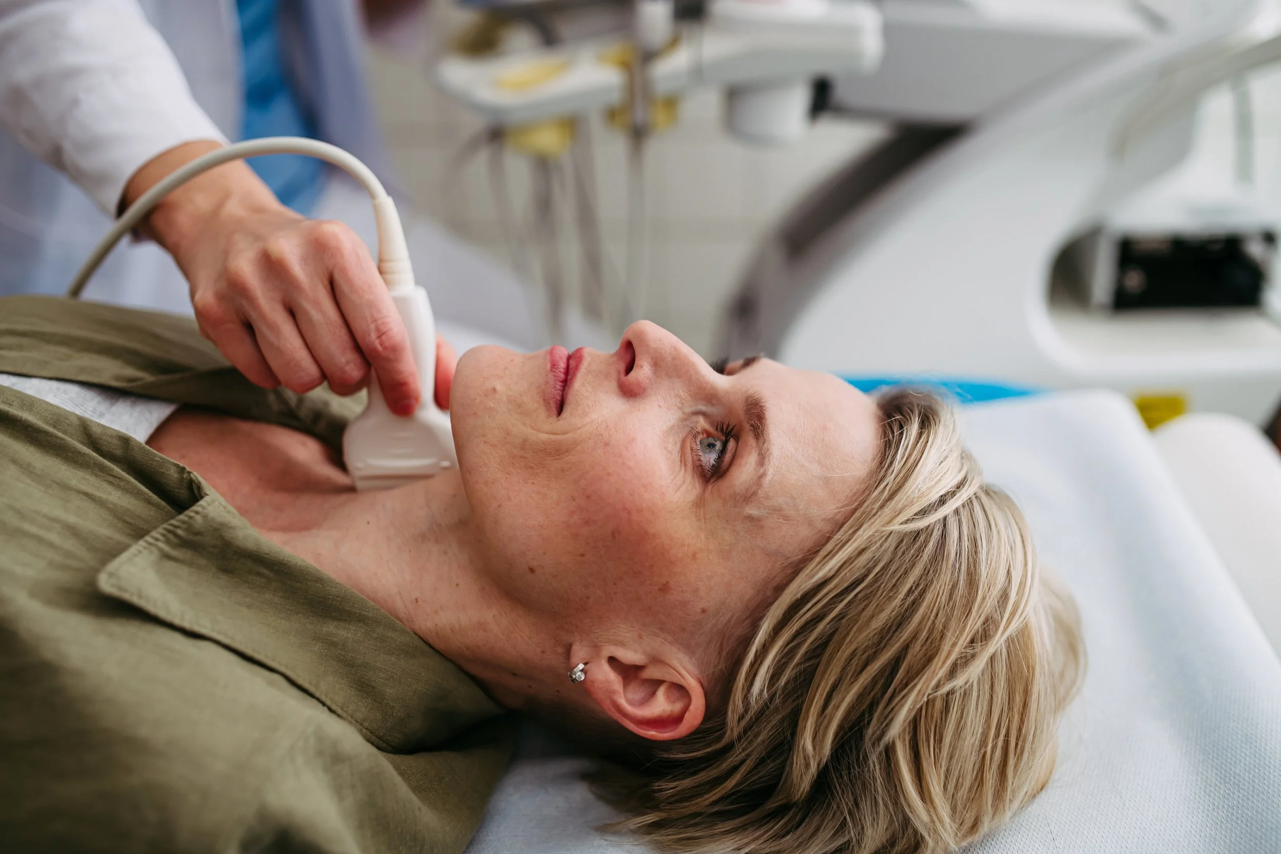 A woman lying on a hospital bed receiving an ultrasound scan of her neck from a medical professional.