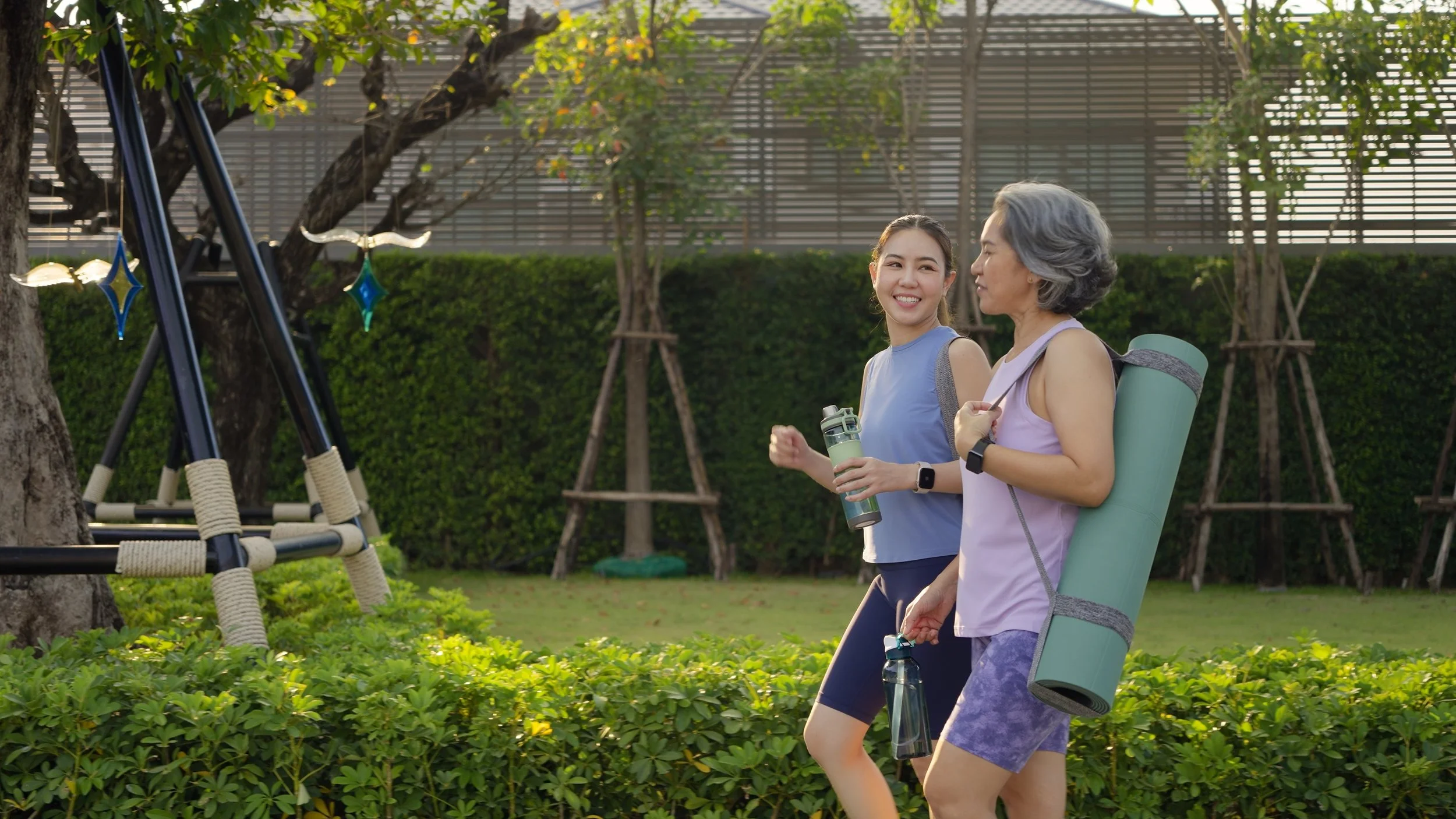 Two women exercising outdoors in a park. One is young, smiling, holding a water bottle, wearing a purple top and dark shorts. The other has gray hair, carrying a yoga mat on her back, wearing a light purple top and purple shorts.