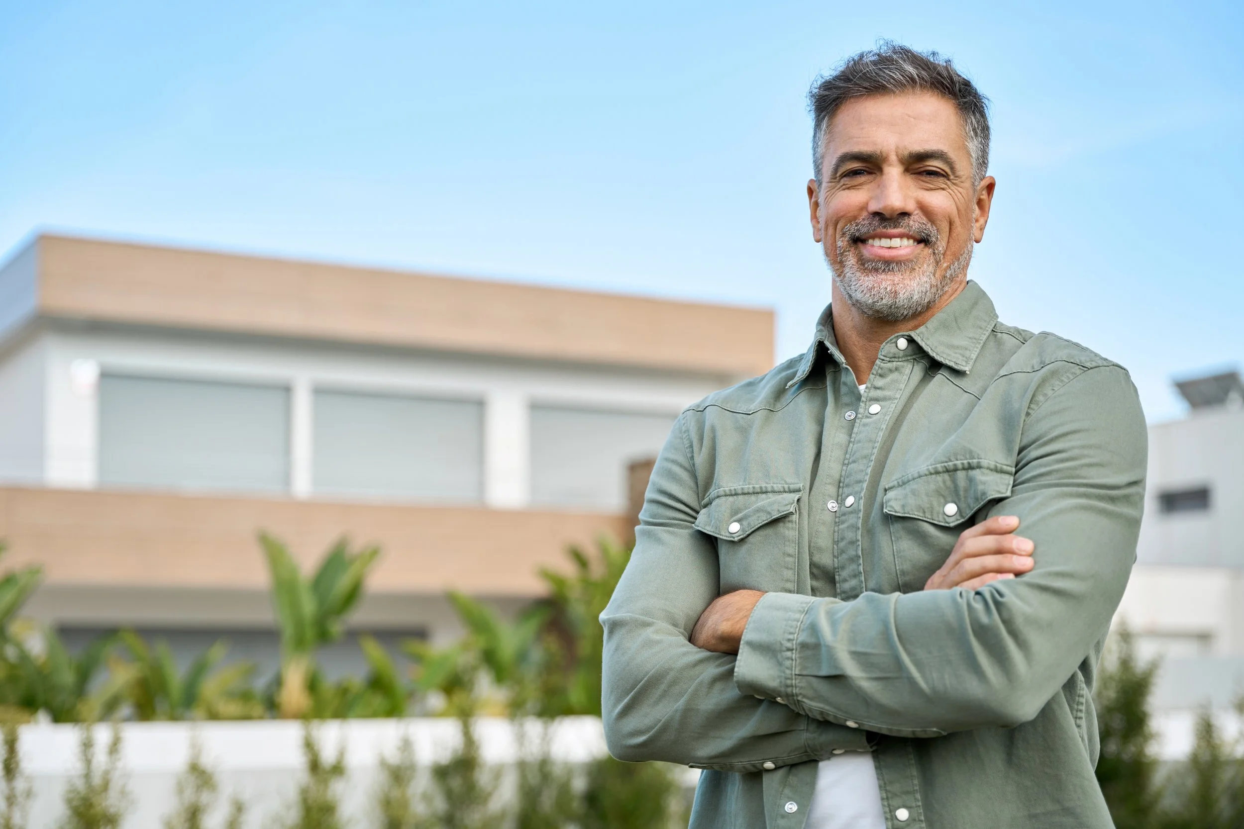 A smiling middle-aged man with gray hair and beard, wearing a green button-up shirt, standing outdoors with modern house and blue sky in the background.