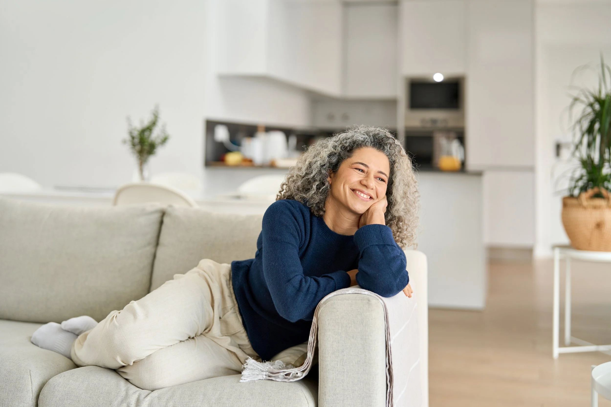 A woman with curly gray hair leaning on a beige sofa armrest, smiling with her head resting on her hand, in a bright, modern living room.