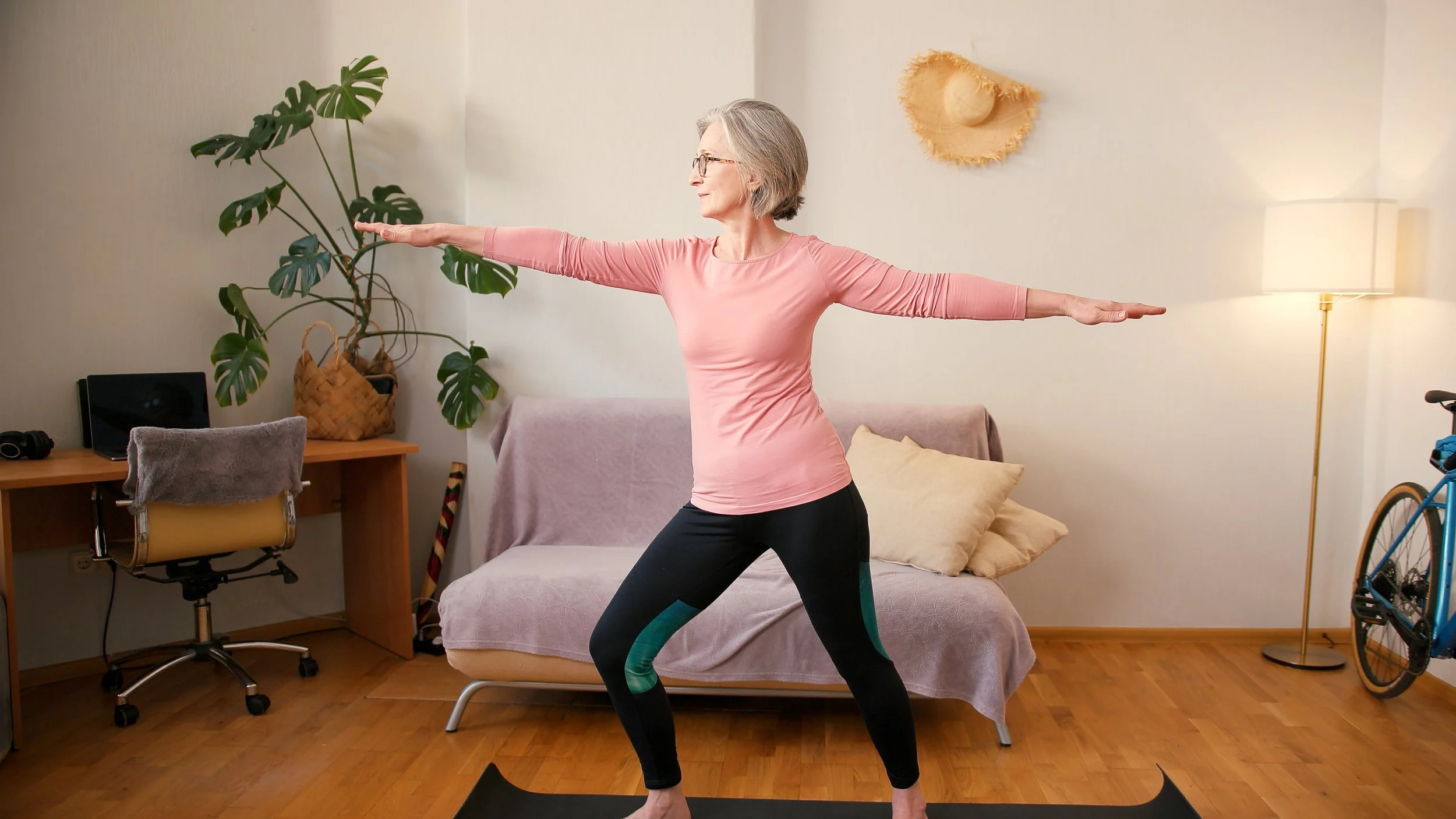 An elderly woman practicing yoga indoors, standing on a black yoga mat with arms extended to the sides, in a living room with a beige couch, wooden floor, large green potted plant, and a bicycle in the background.