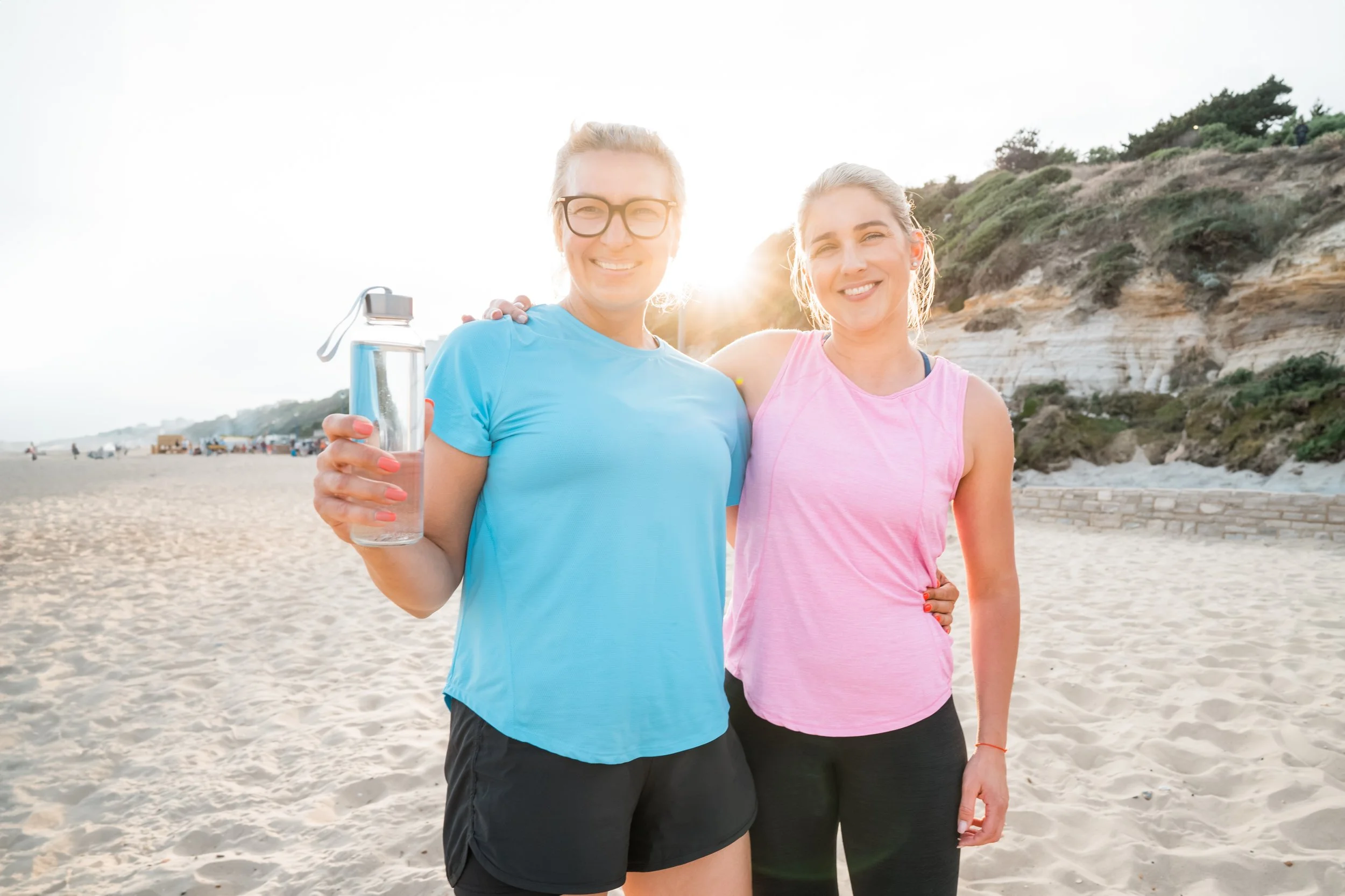 Two women smiling at the beach, one holding a water bottle, with cliffs and beachgoers in the background.