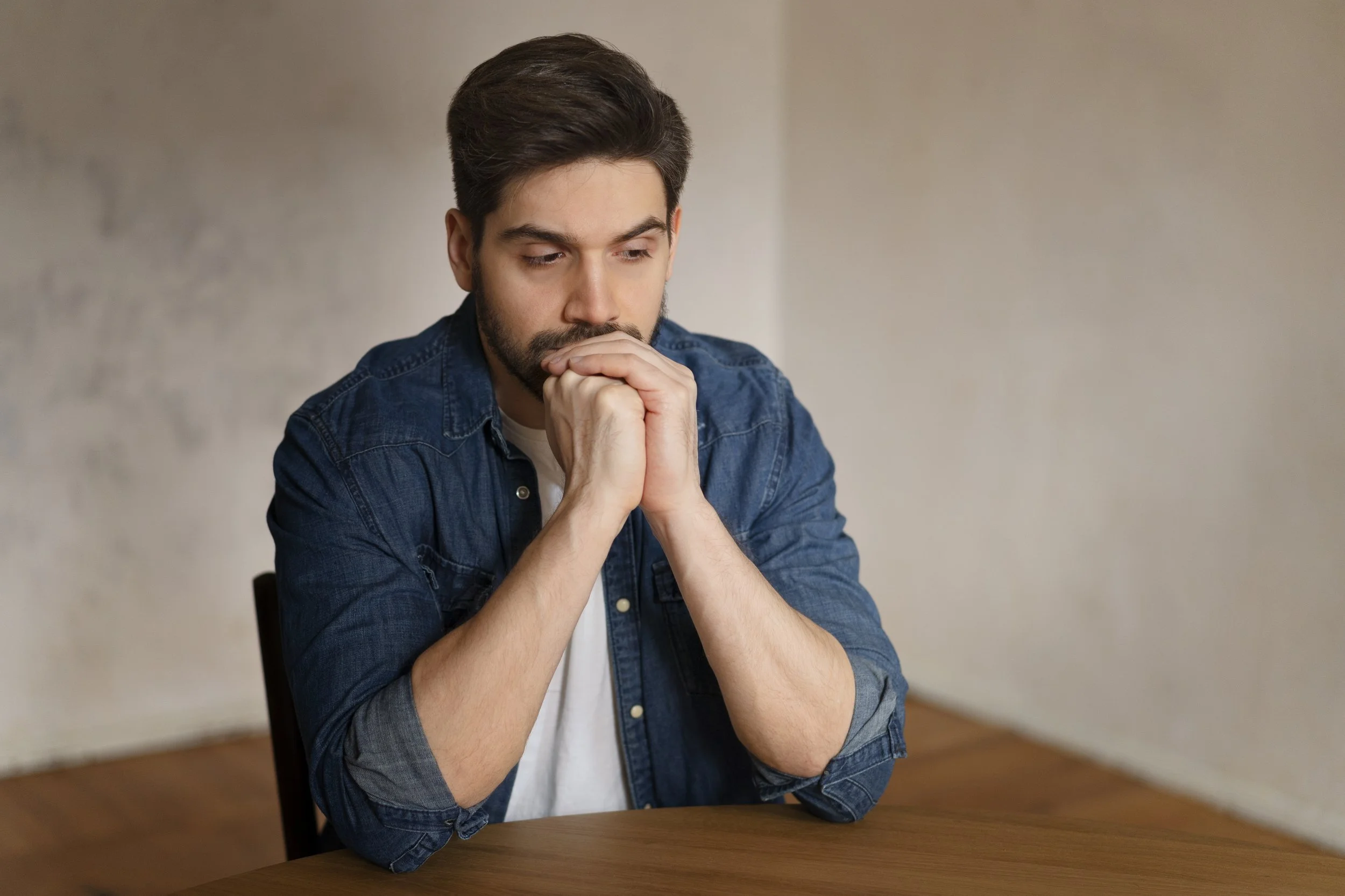 A young man with dark hair and beard, wearing a denim shirt, sitting at a wooden table with hands clasped near his mouth, appeared to be deep in thought or worried.
