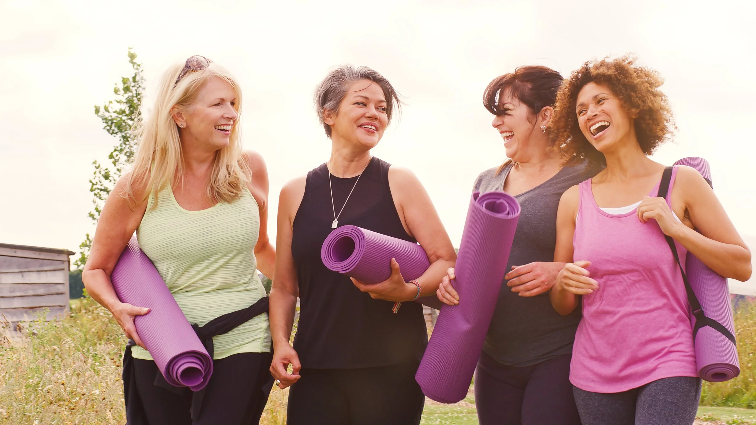 Four women standing outdoors with yoga mats, smiling and chatting.