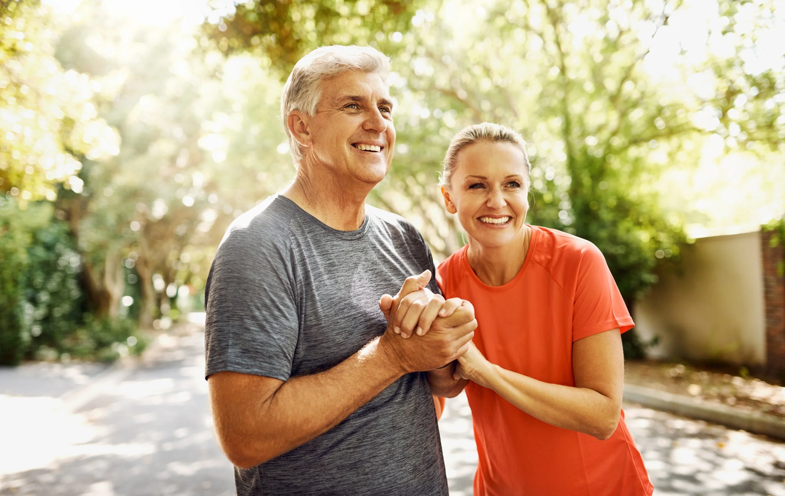 A happy elderly man and woman holding hands and smiling outdoors on a sunny day, surrounded by green trees.
