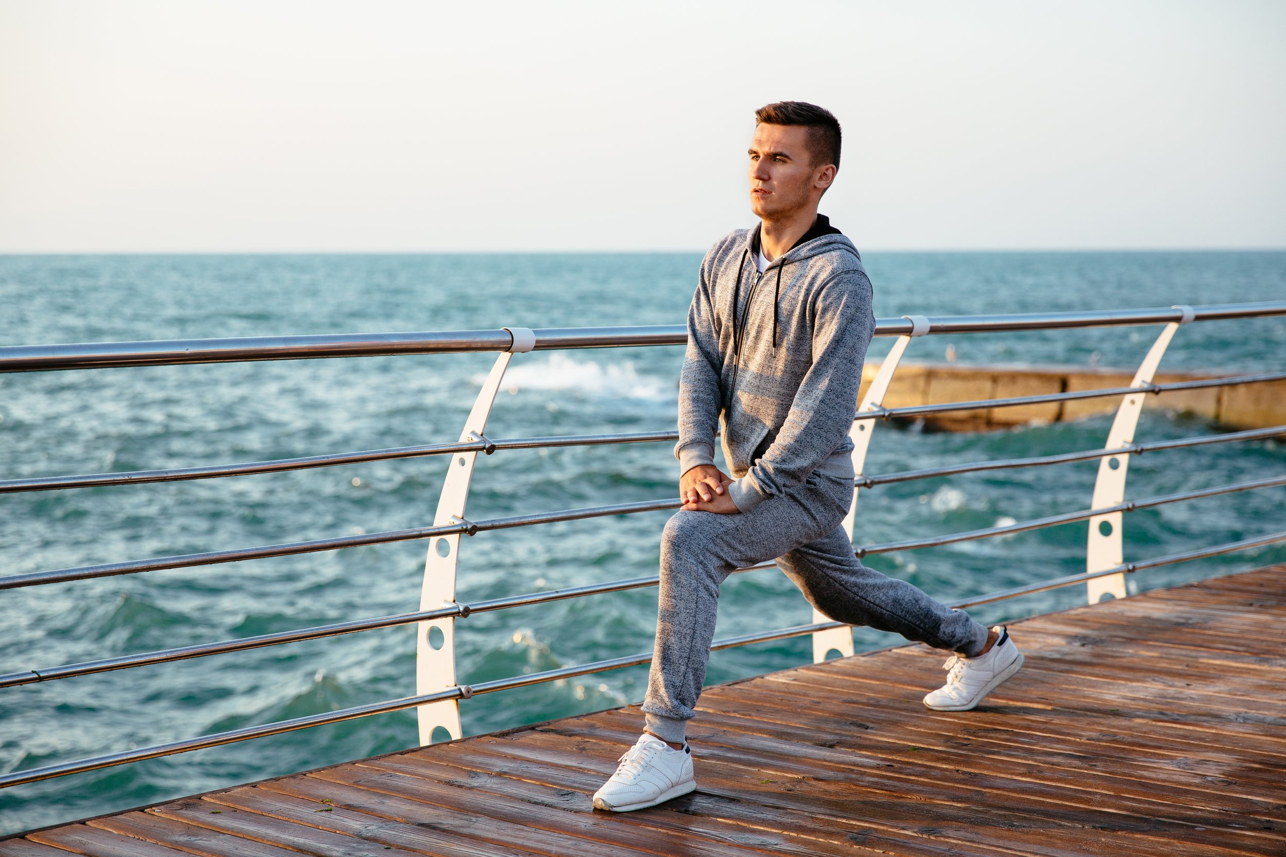A man in athletic gray hoodie and sweatpants doing a stretch on a wooden pier by the ocean during sunset.