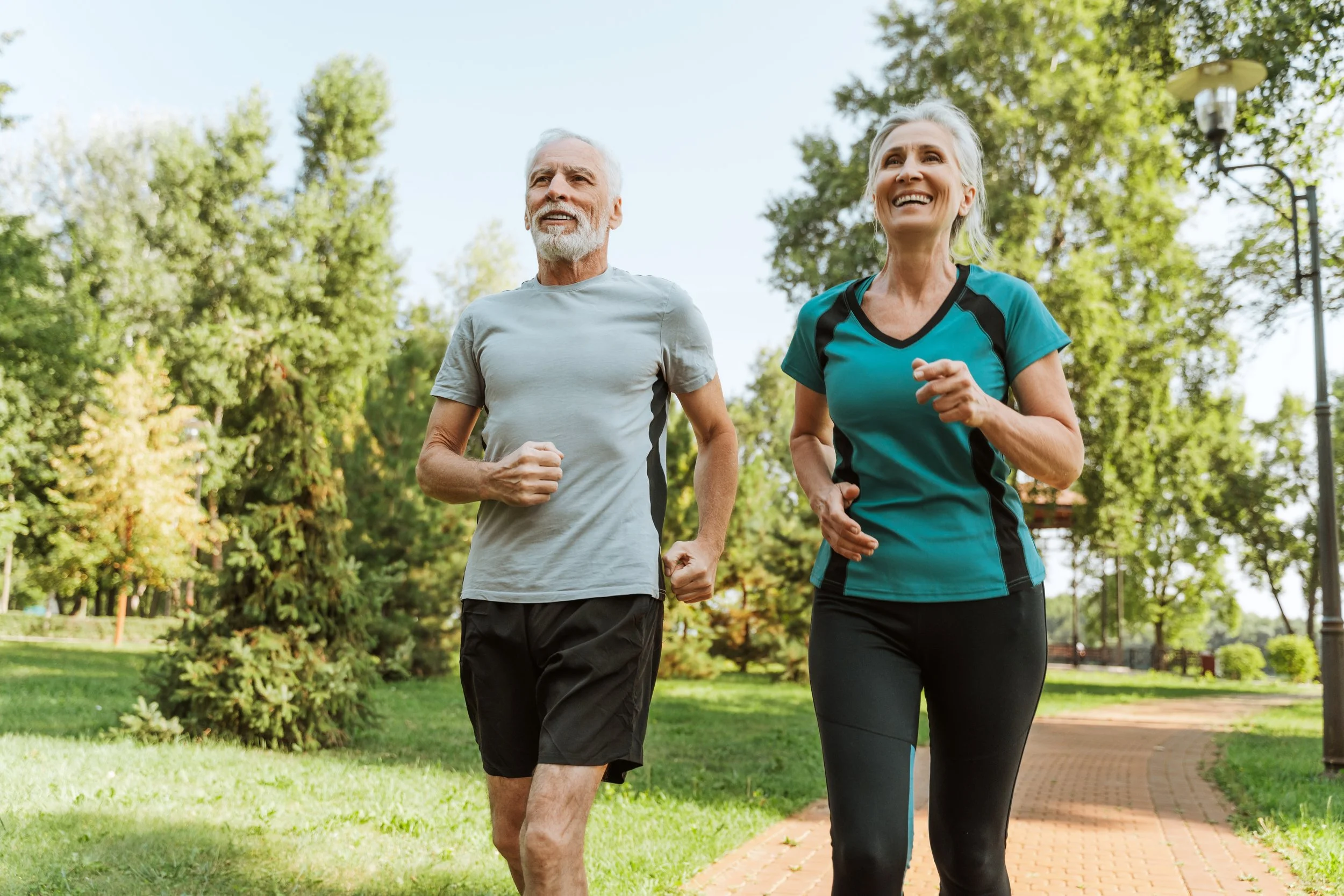 An elderly man and woman jogging together in a park on a sunny day, surrounded by green trees.