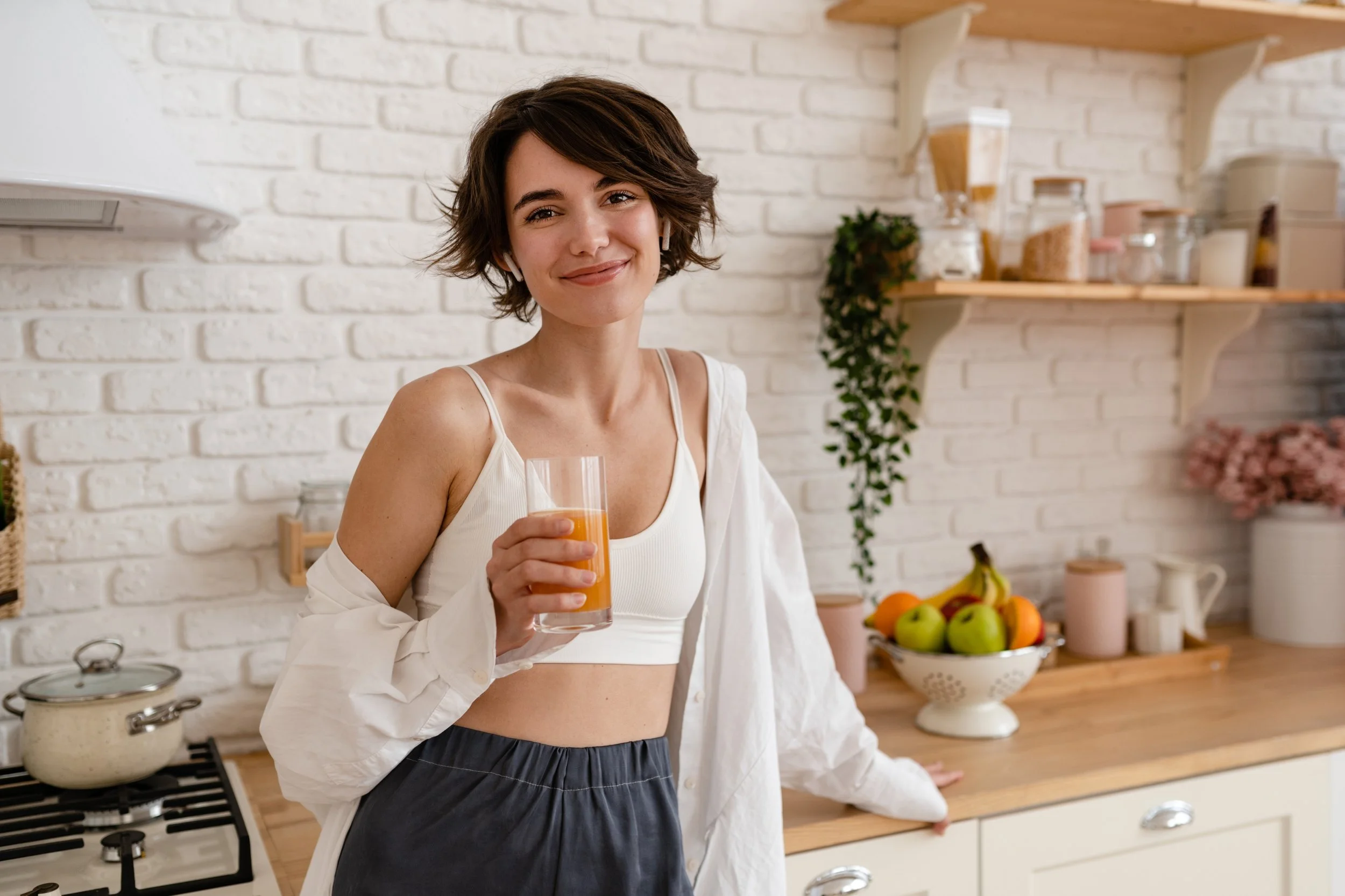 A woman with short brown hair, wearing a white tank top and dark pants, is standing in a kitchen holding a glass of orange juice, smiling.