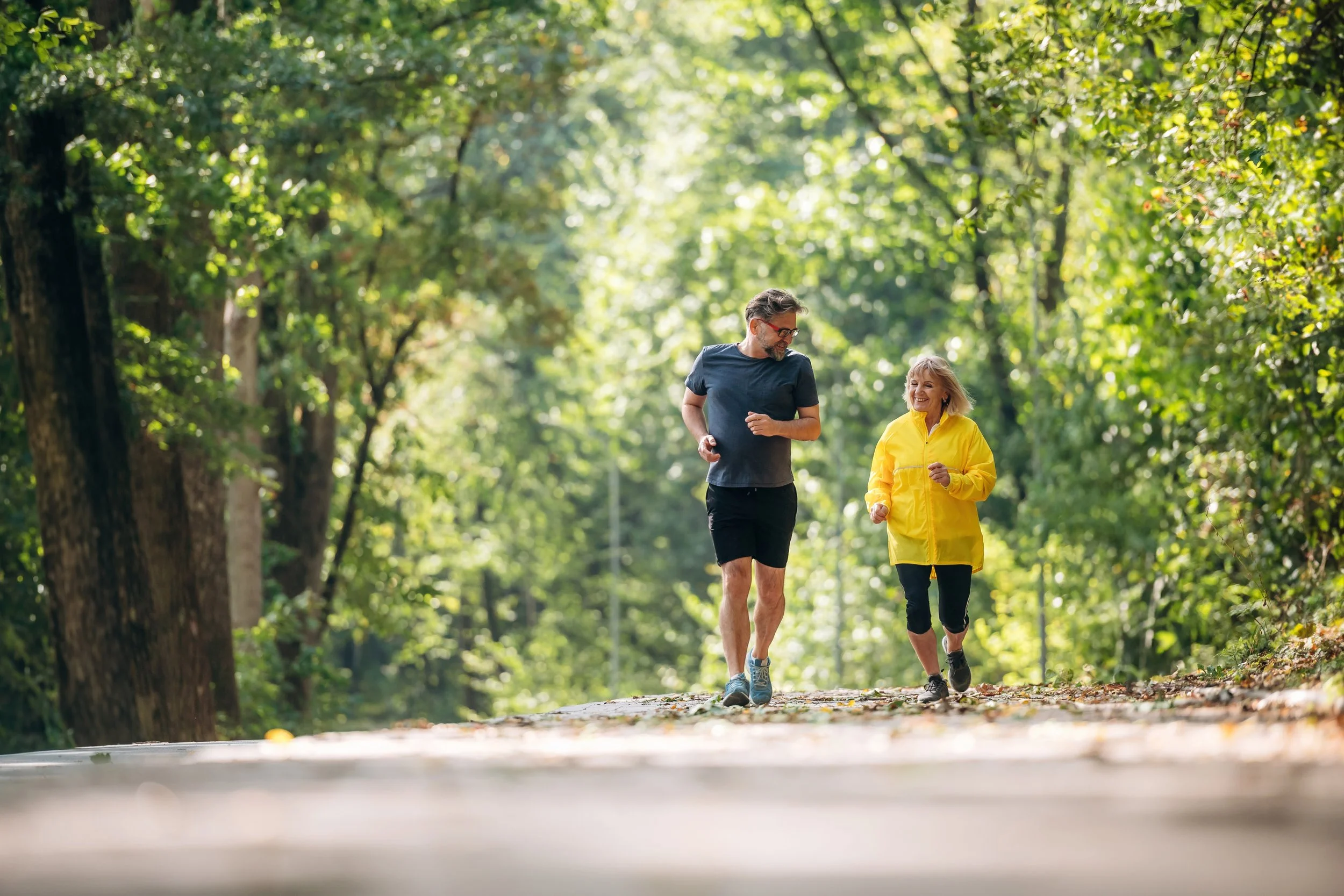 A man and woman jogging together on a forest trail surrounded by lush green trees, enjoying a sunny day.