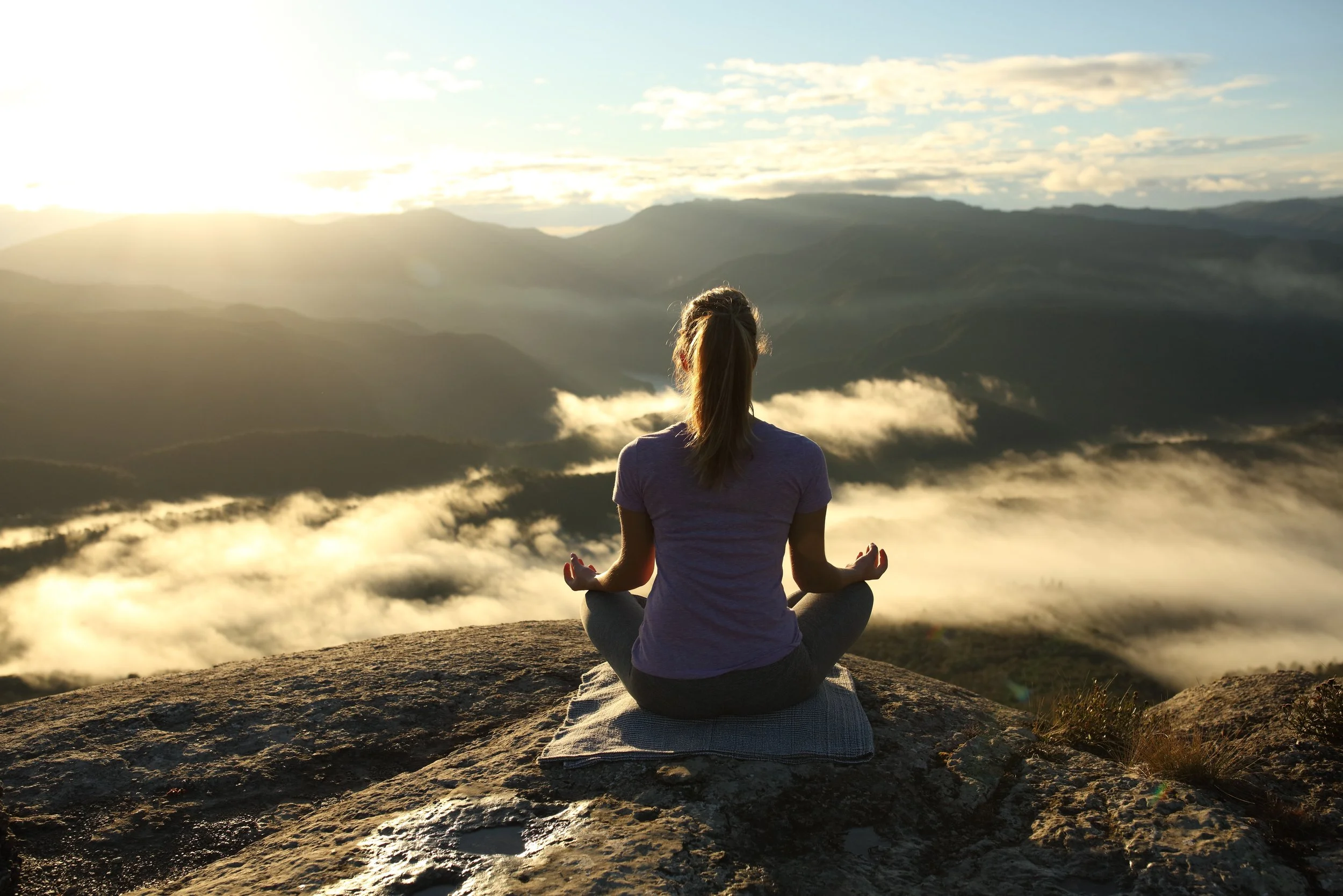 A woman practicing yoga on a rock in a meditative pose, facing a mountain landscape at sunrise with clouds and sunlight.