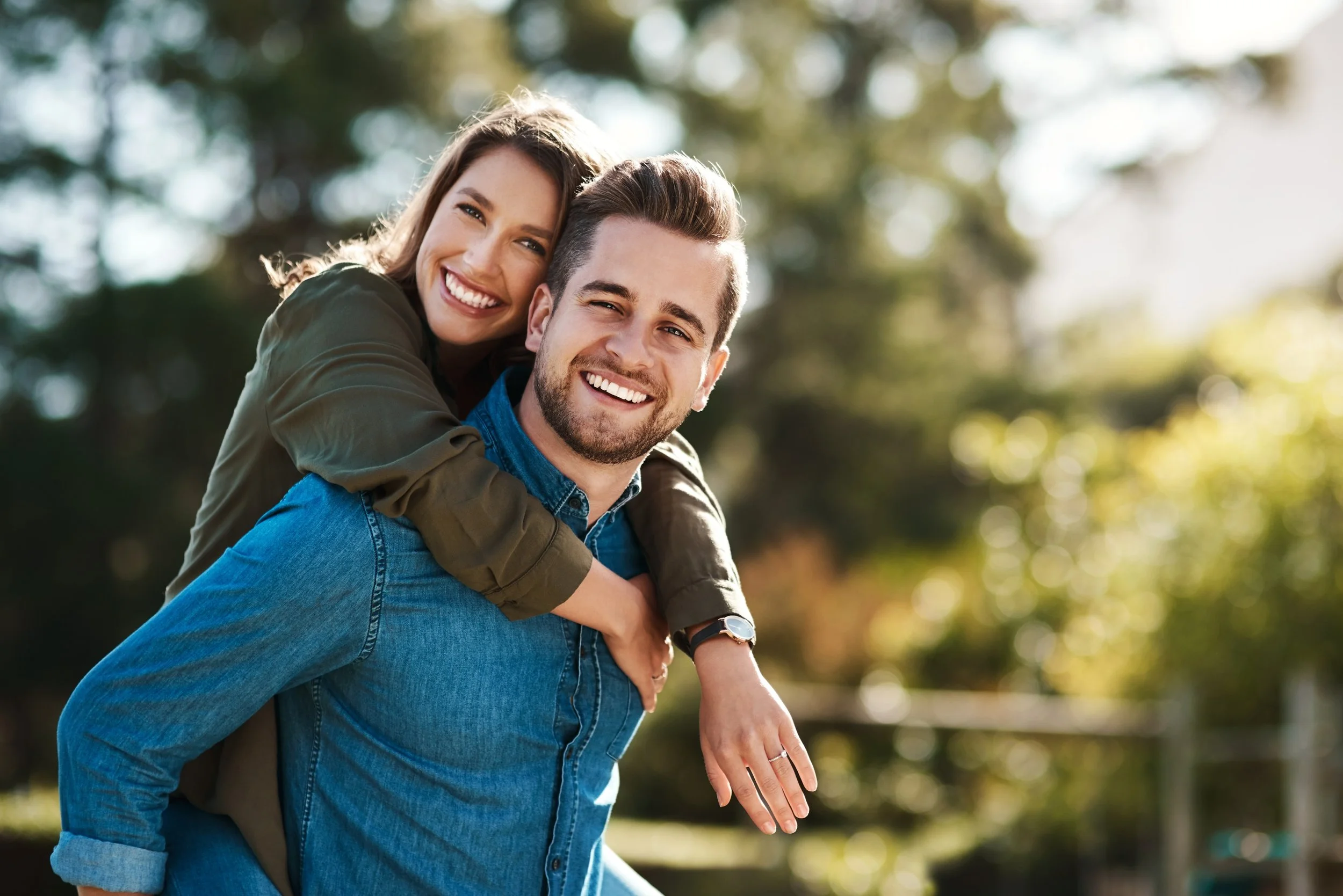A happy couple outdoors, with the woman giving the man a piggyback ride, both smiling at the camera, with trees and sunlight in the background.