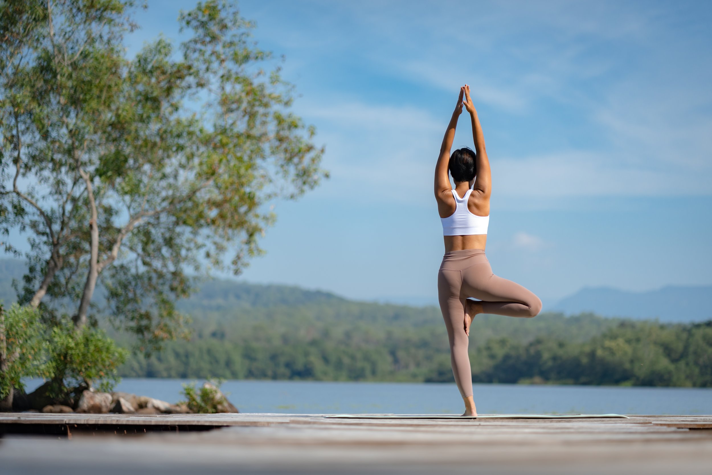 A woman practicing yoga outdoors on a dock near a lake, standing in tree pose with arms raised above her head against a backdrop of blue sky, trees, and distant mountains.