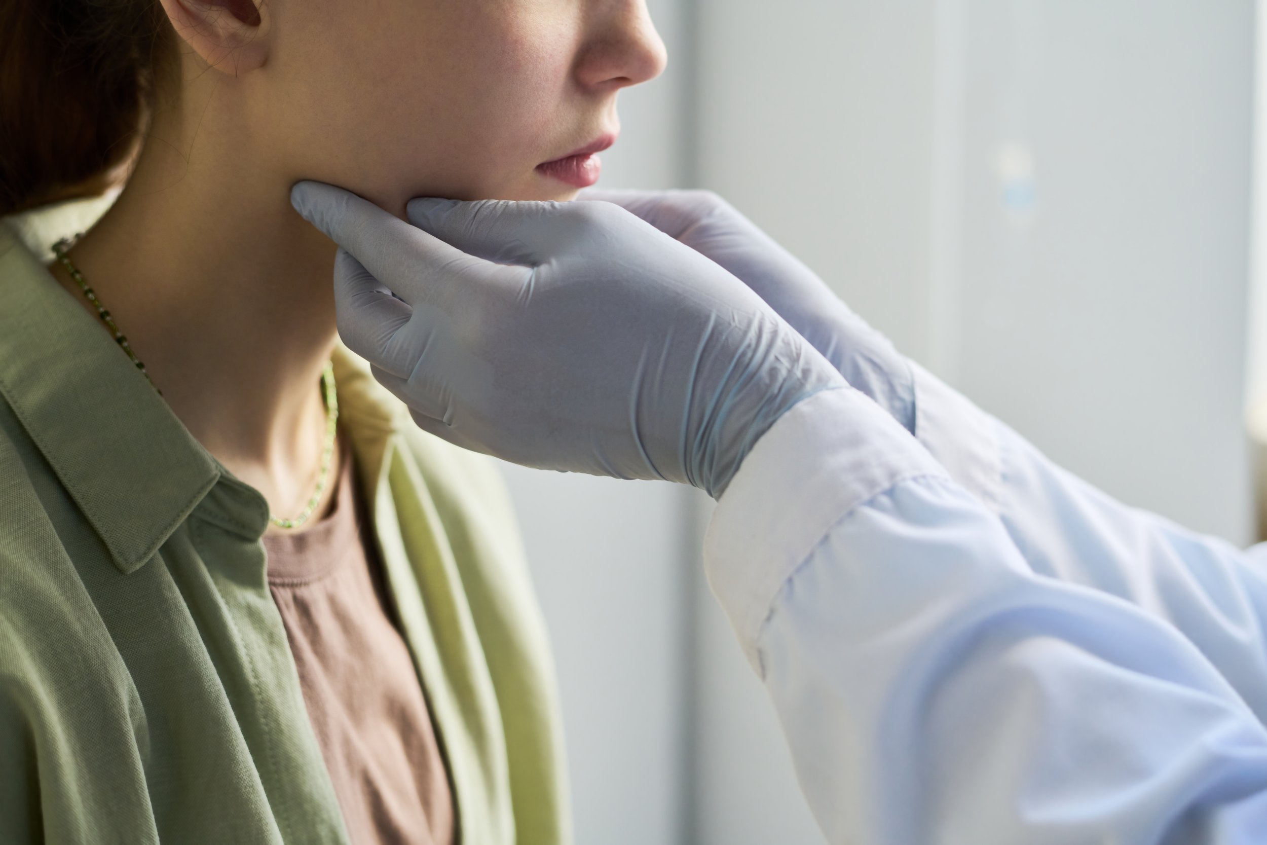 A healthcare professional in a white coat with gloved hands gently examines a young woman's chin and neck area.