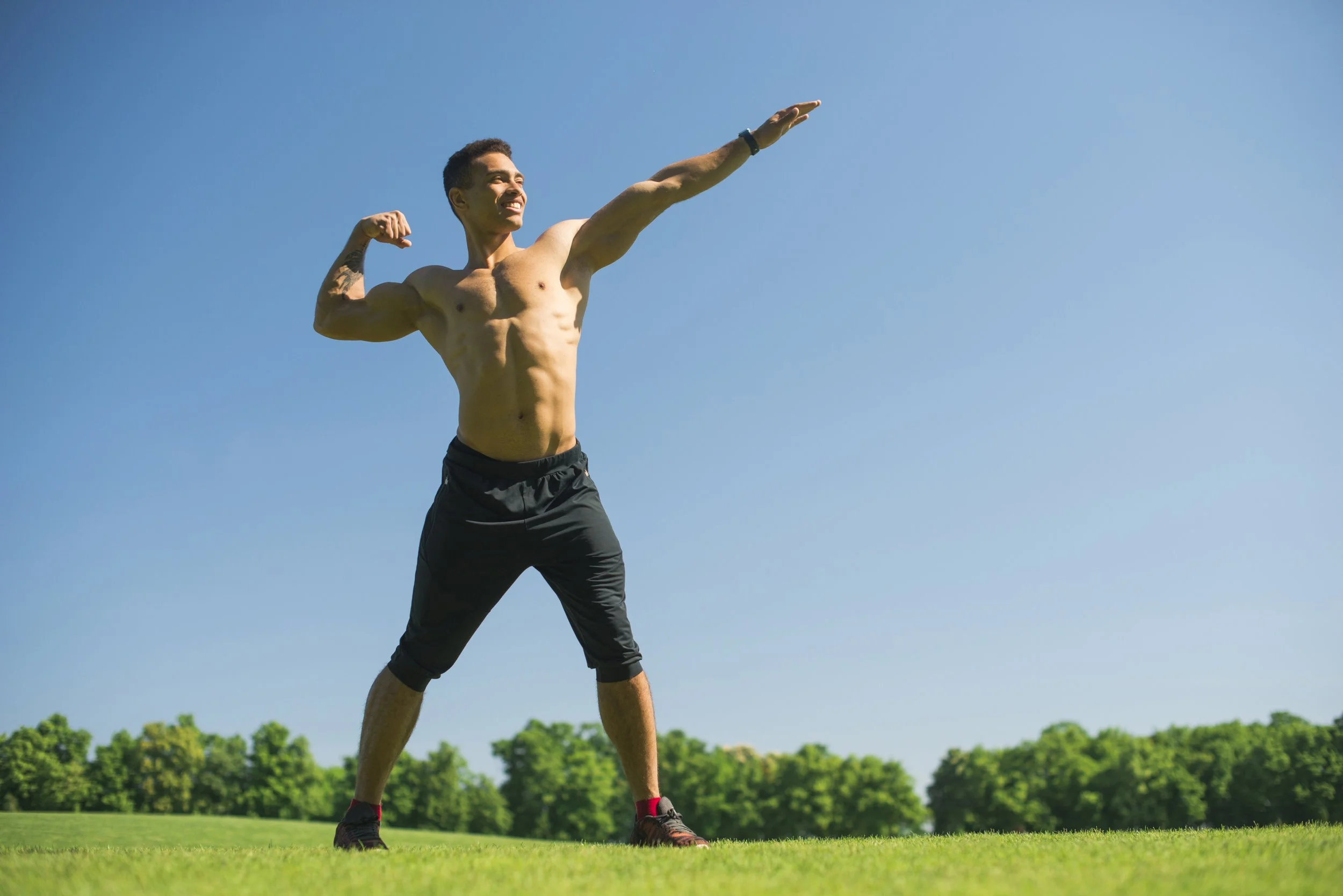 A fit, shirtless young man flexing his right arm and extending his left arm outdoors on a sunny day, with a grassy field and trees in the background.