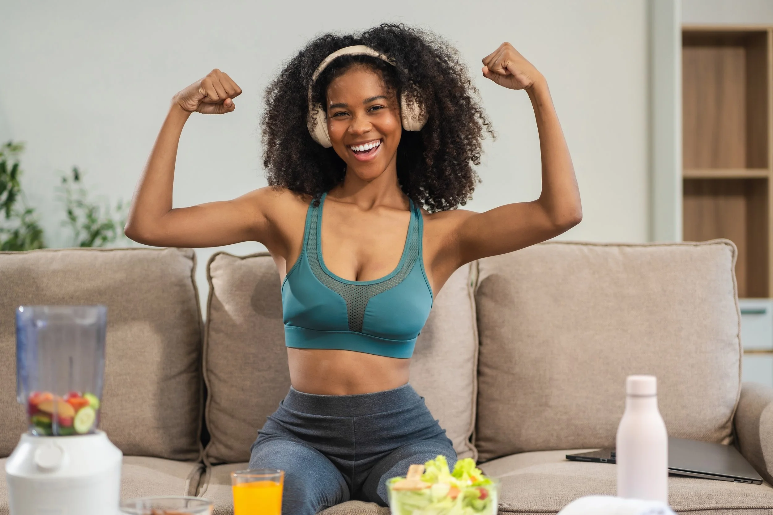 A woman sitting on a beige couch, flexing her biceps, smiling, wearing a teal sports bra and headphones, with a blender, a glass of orange juice, a bottle, a laptop, and a salad on the coffee table in front of her.