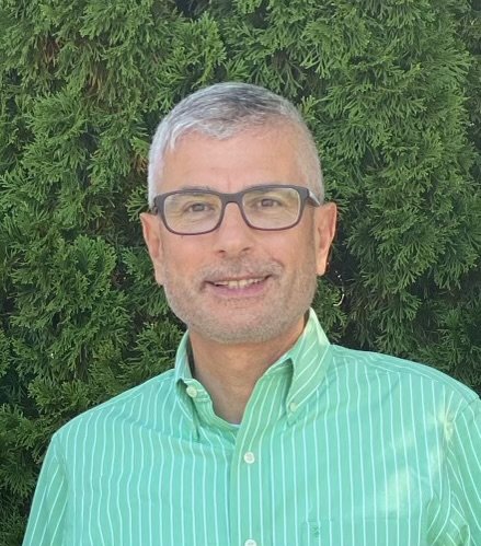 Close-up of a middle-aged man with gray hair, glasses, smiling, outdoors with greenery and brick building in background.