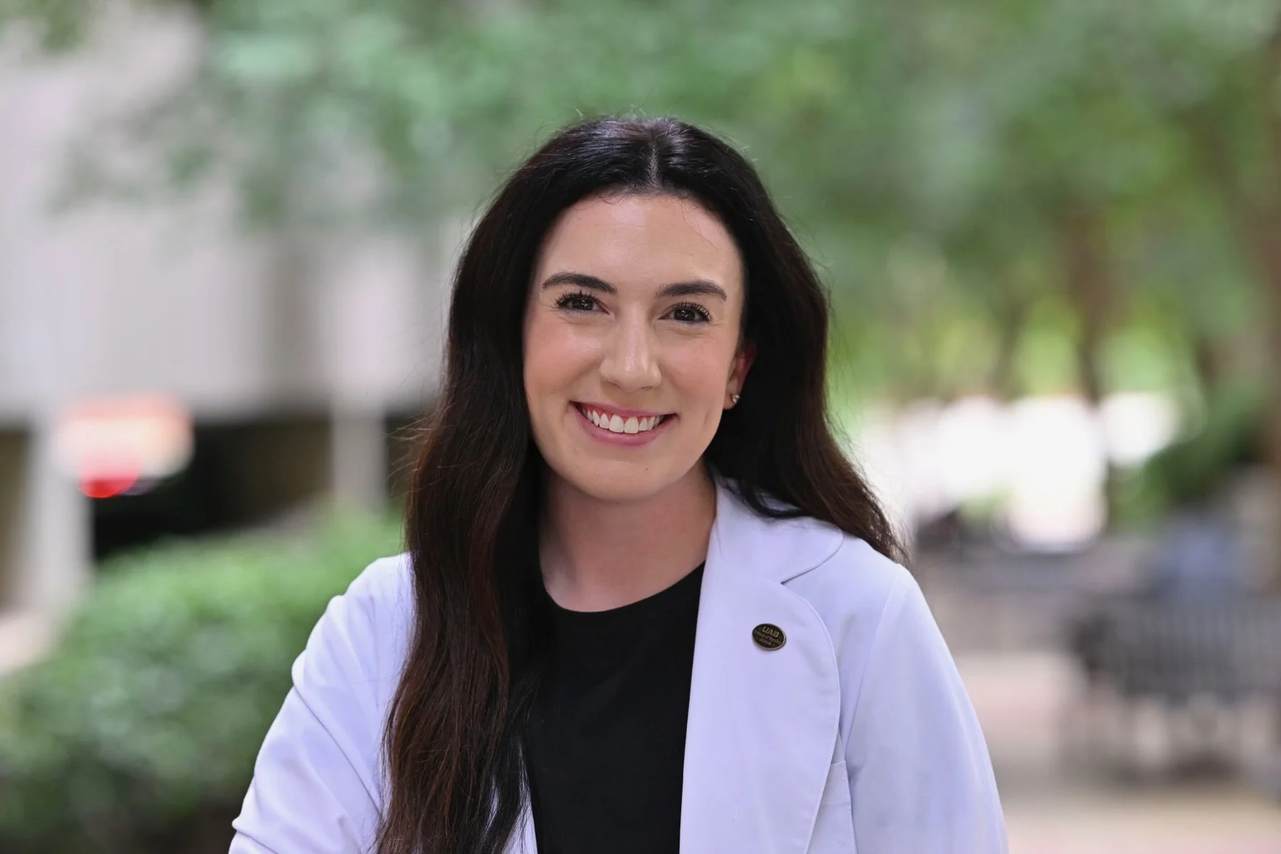 A young woman with long dark hair, smiling, wearing a white jacket over a black top, standing outdoors with green trees and blurred background.