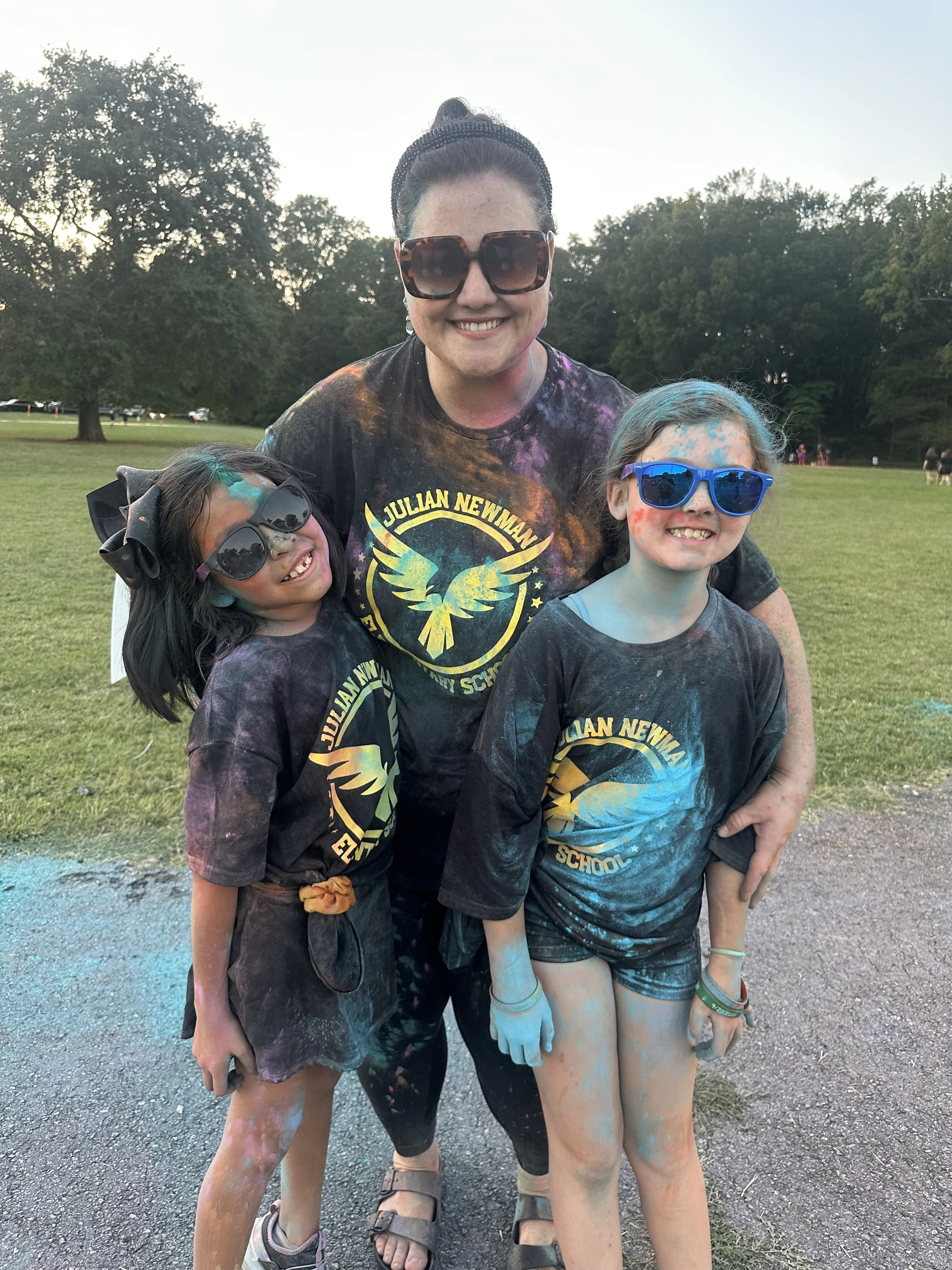 A woman and two young girls at a color run event, all smiling and wearing sunglasses. They are covered in colorful powder and wearing matching black t-shirts with a logo for Julian Newman Elementary School, outdoors on a grassy field with trees in the background.