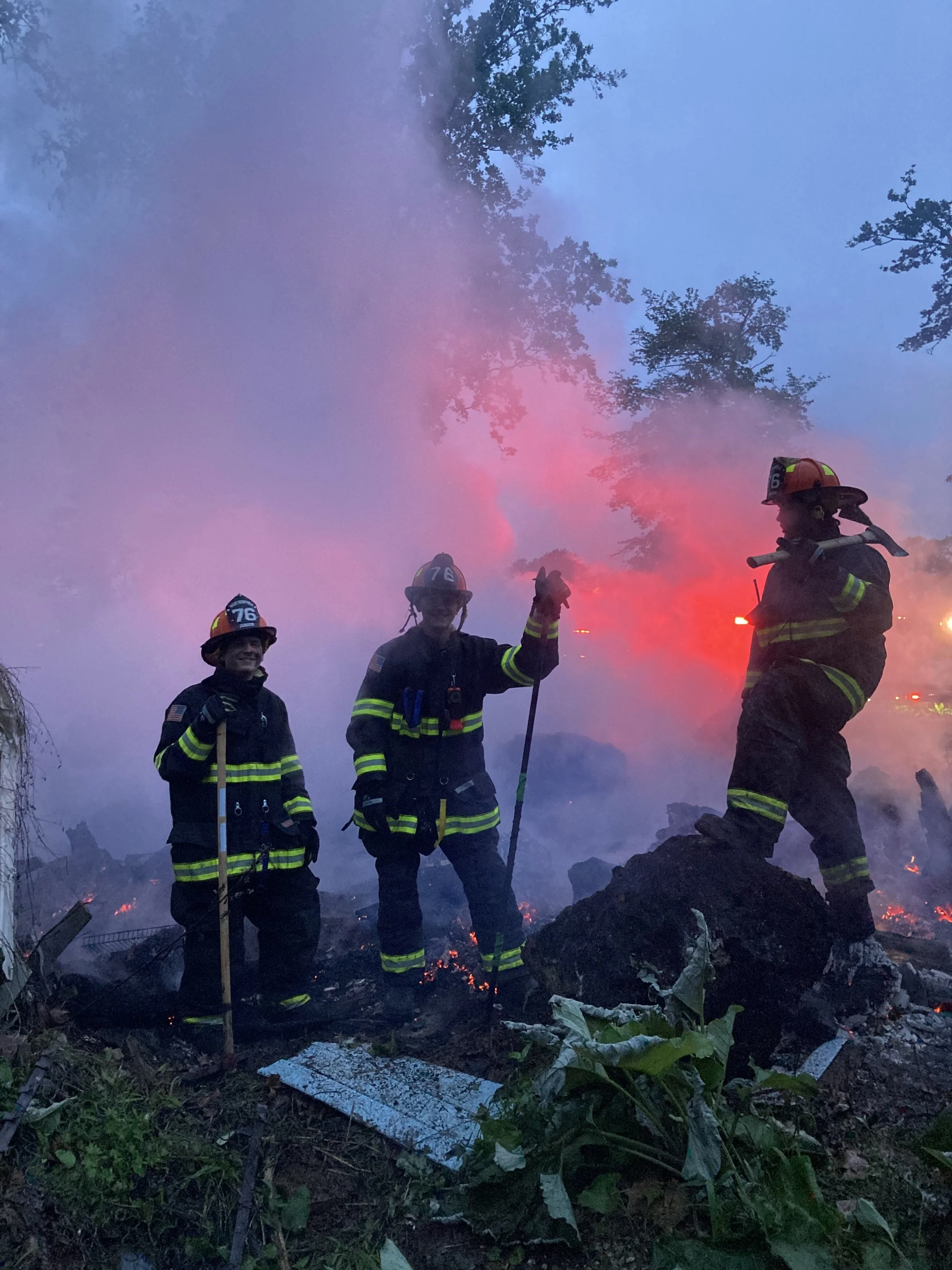 Three firefighters in full gear standing amidst the debris of a fire, with smoke and flames in the background. The scene appears to be at dusk or dawn.