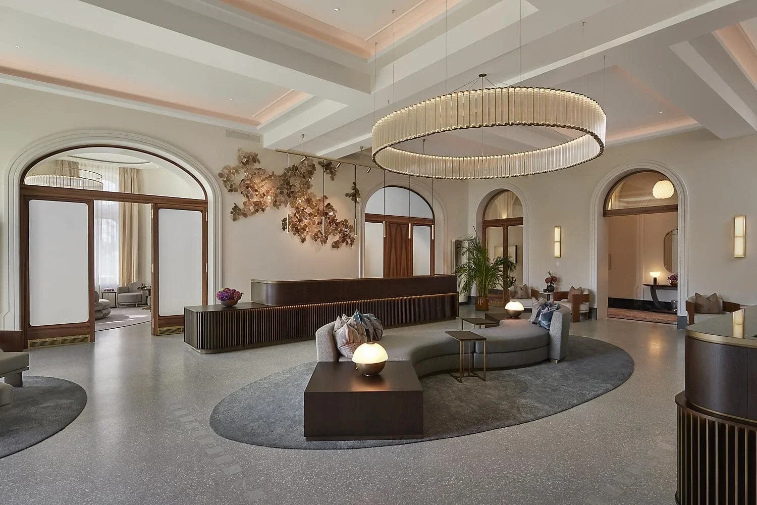 Luxury hotel lobby with modern decor, curved seating, a large ring chandelier, and neutral tones.