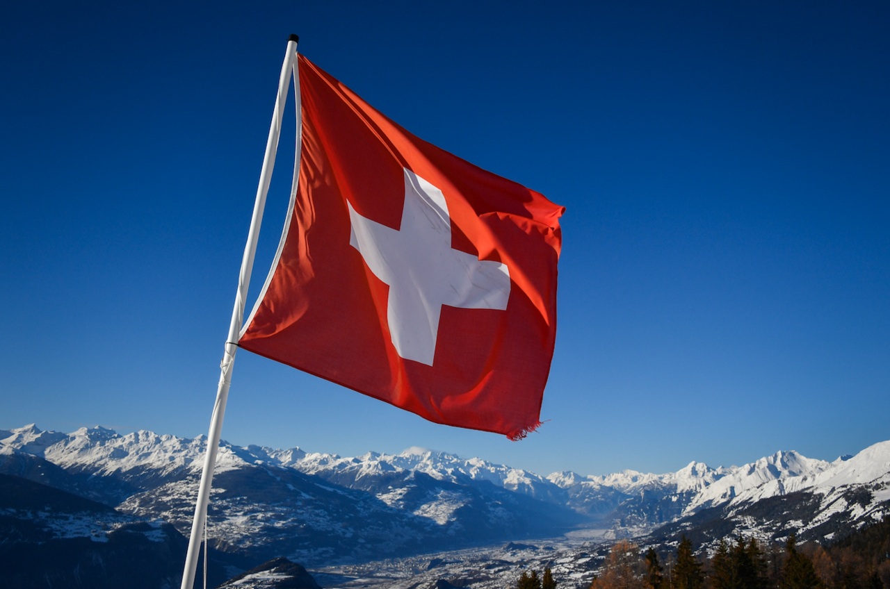 Switzerland flag against a mountain landscape with snowy peaks and a clear blue sky.