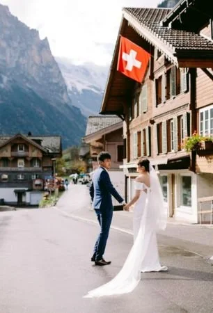 A wedding couple holding hands in a mountain village street with Swiss flags, traditional buildings, and mountains in the background.