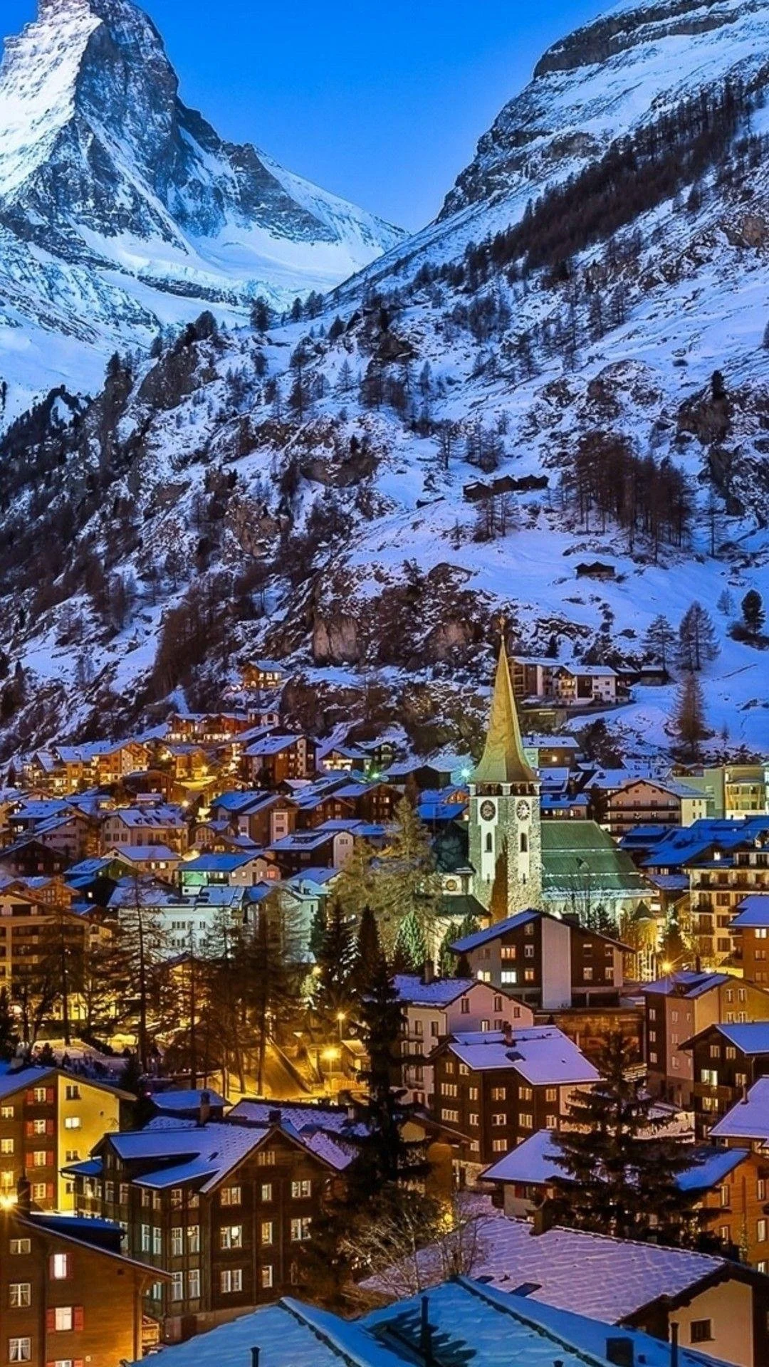 A snow-covered mountain village at dusk with illuminated buildings and a prominent clock tower church, surrounded by pine trees and snow-capped mountains in the background.