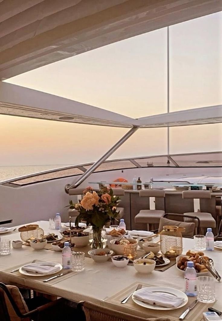 A dining table set with plates, glasses, and various bowls of food on a yacht, with a view of the ocean and sunset in the background.