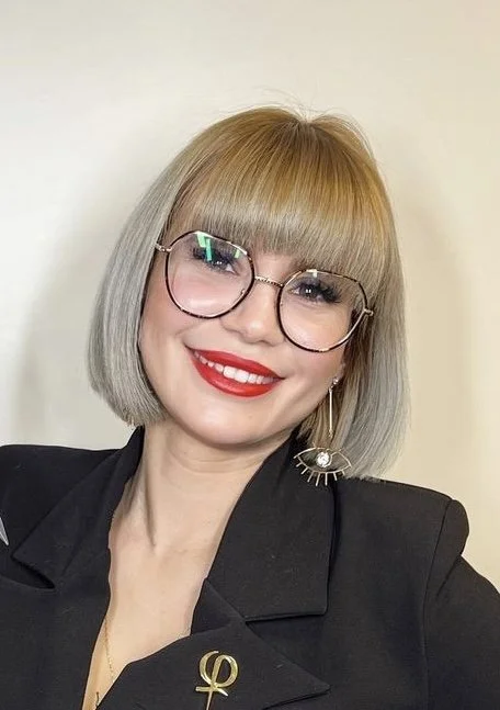 A woman with blonde hair and bangs, wearing glasses and bright red lipstick, smiling at the camera. She has a black blazer with a gold omega symbol on the lapel, and a large eye-shaped earring on her left ear.