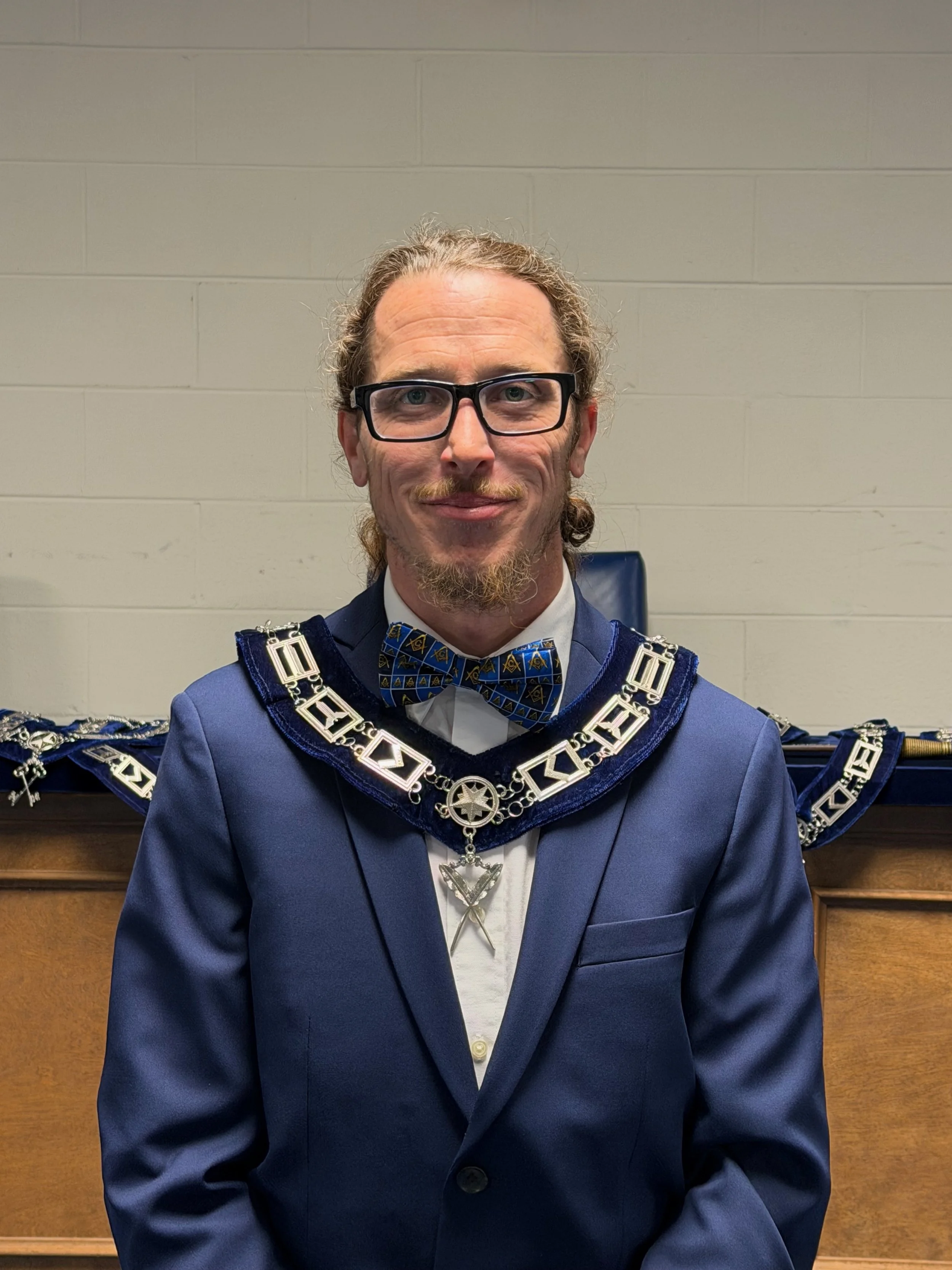 A man with glasses, long curly hair, and a beard wearing a blue suit with a bow tie, and a ceremonial chain around his neck stands in front of a wooden background.