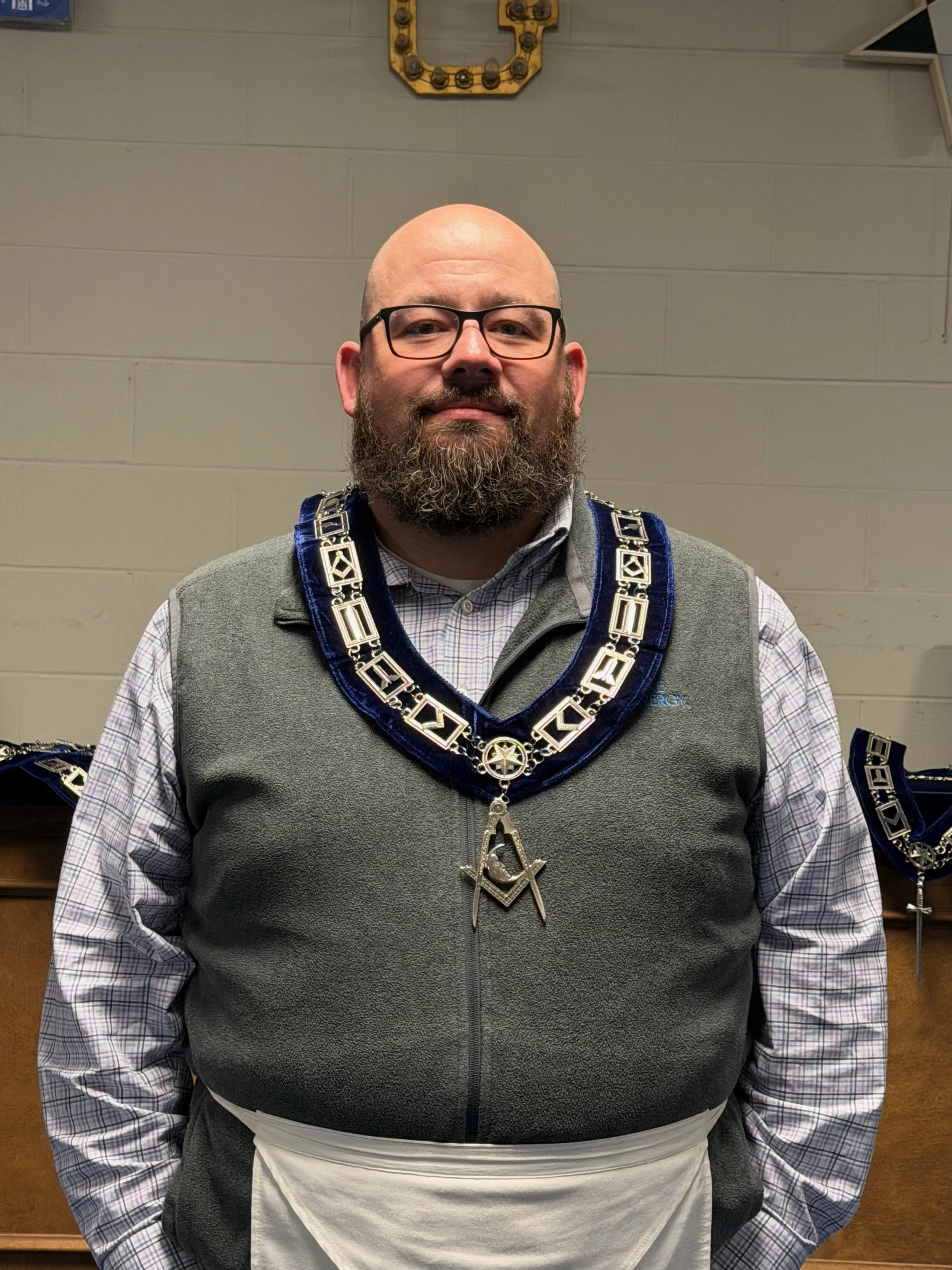 A man with glasses and a beard wearing Masonic regalia, including a decorated collar and apron, standing indoors against a plain wall.