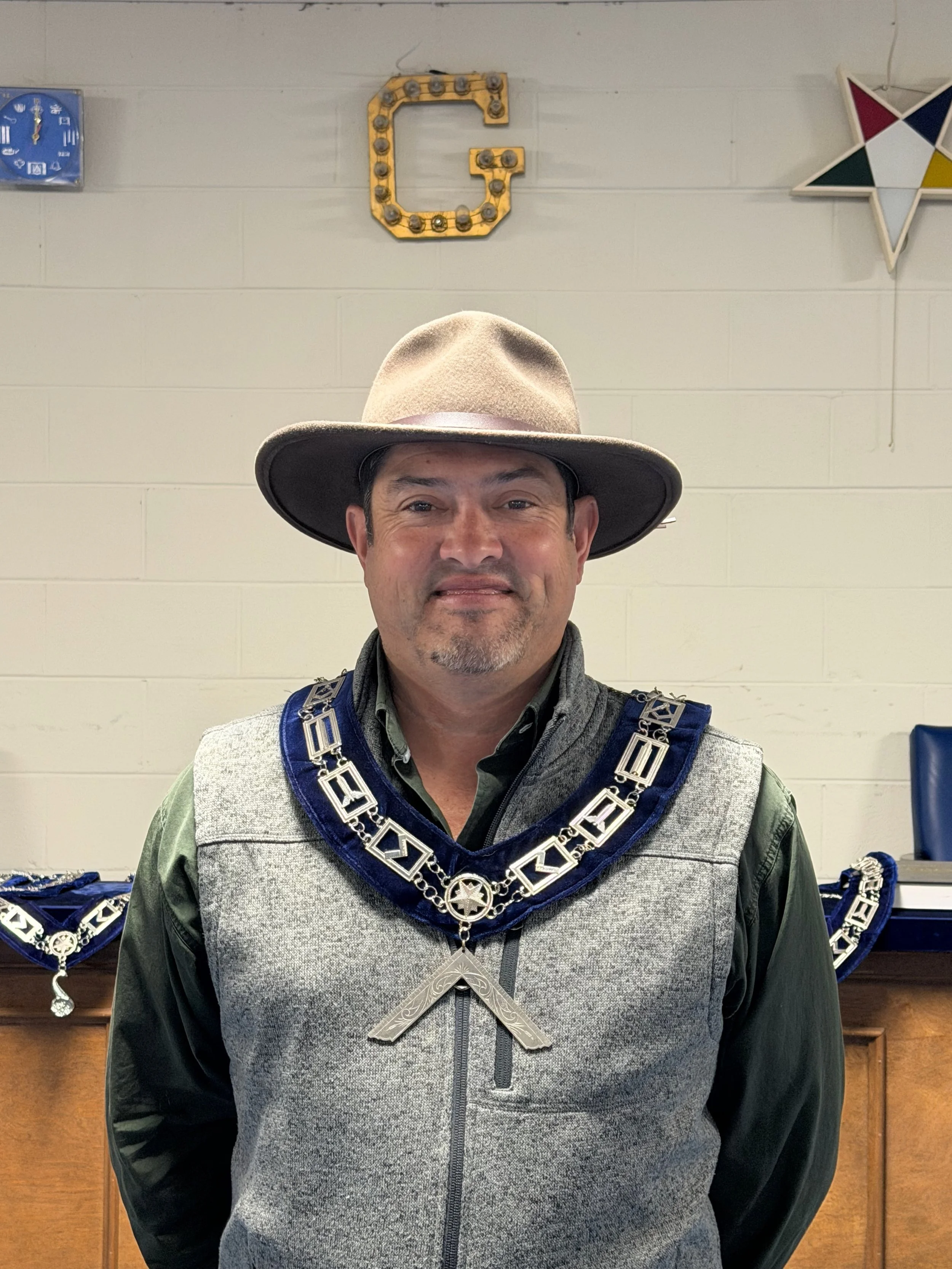 A man wearing a wide-brimmed hat and decorative chain necklace stands in front of a wall decorated with a large letter 'G' and part of a star-shaped wall decoration.
