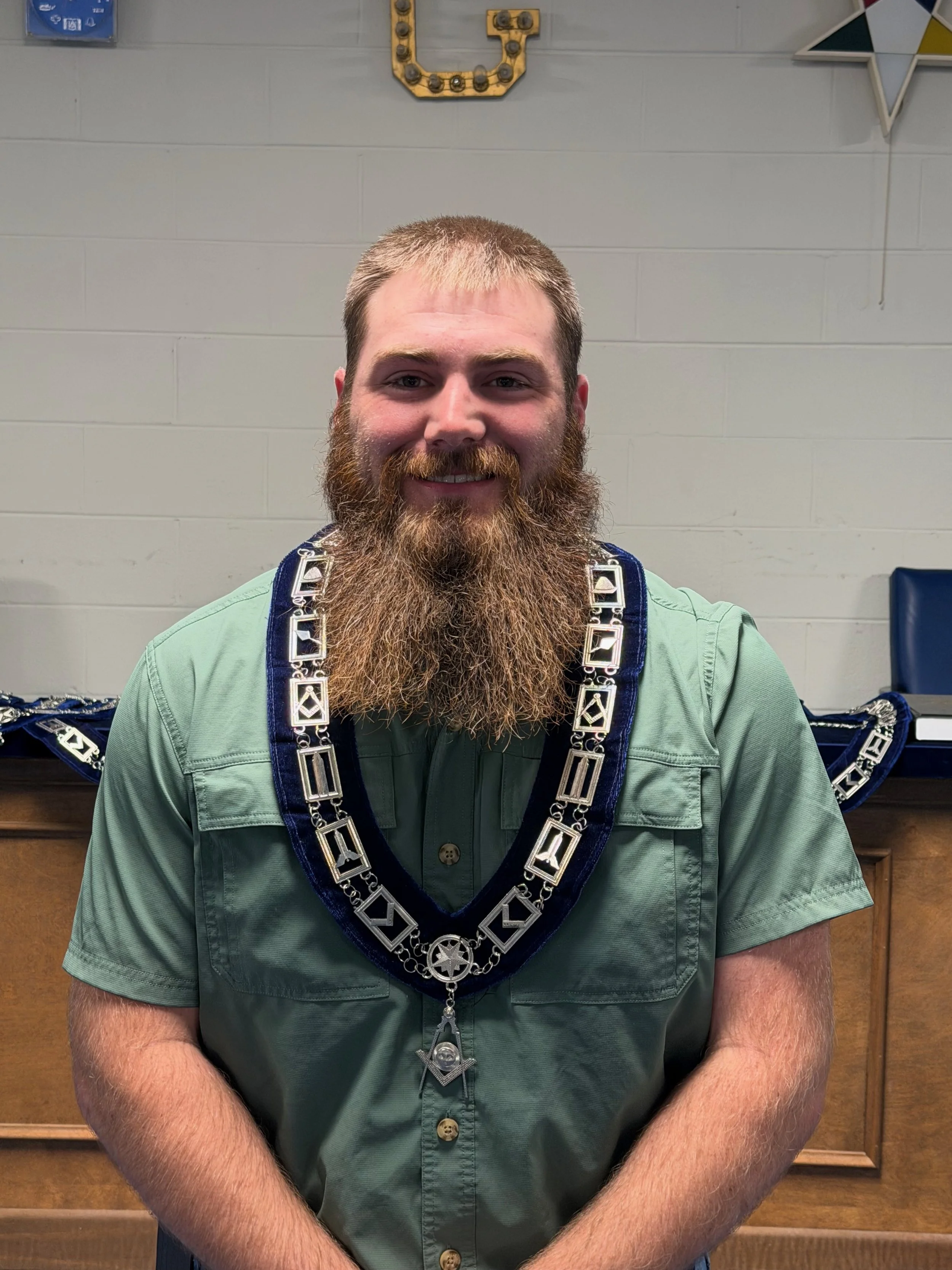 A man with a beard smiling, wearing Masonic regalia including a chain collar with square and compass symbols, in an indoor setting with a wooden cabinet and Masonic decorations in the background.