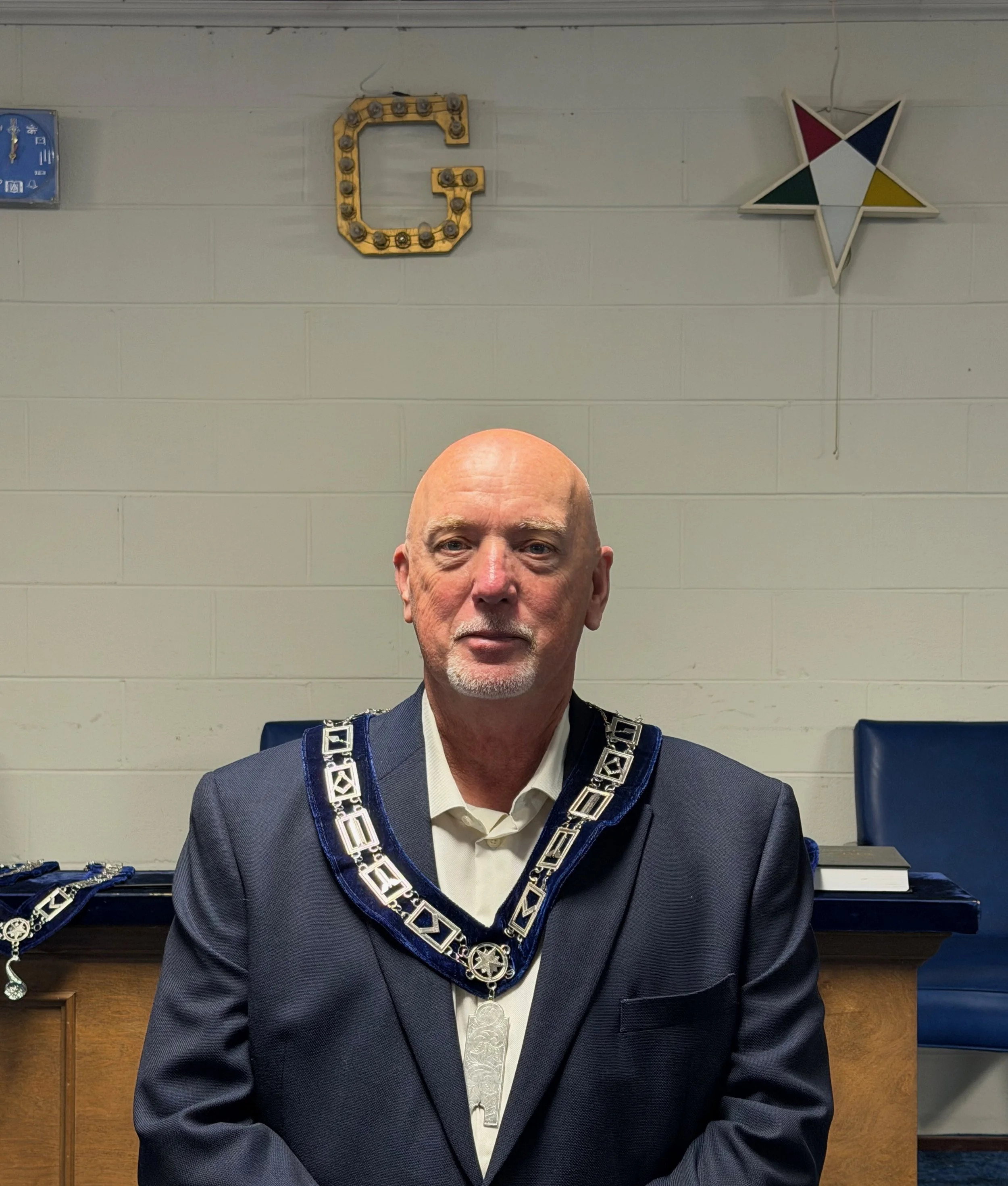 A man in a suit and ceremonial collar sitting in a room with a plain wall, a book on the table, and decorative symbols on the wall including a large letter 'G' with lights and a star-shaped symbol.