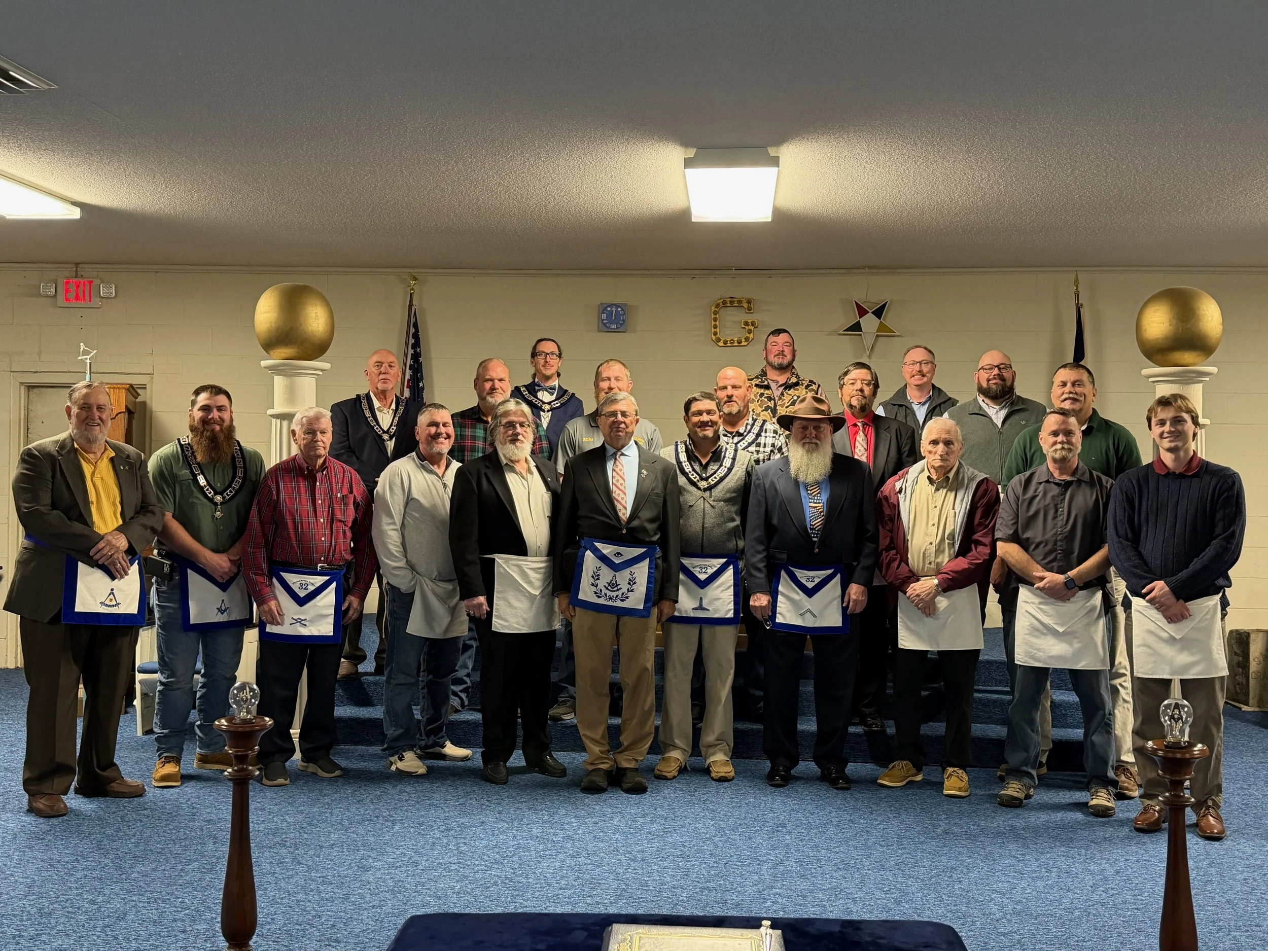A group of men in suits and casual clothing standing in a Masonic lodge, some wearing aprons and regalia, posing for a photo in front of a wall with Masonic symbols and flags.