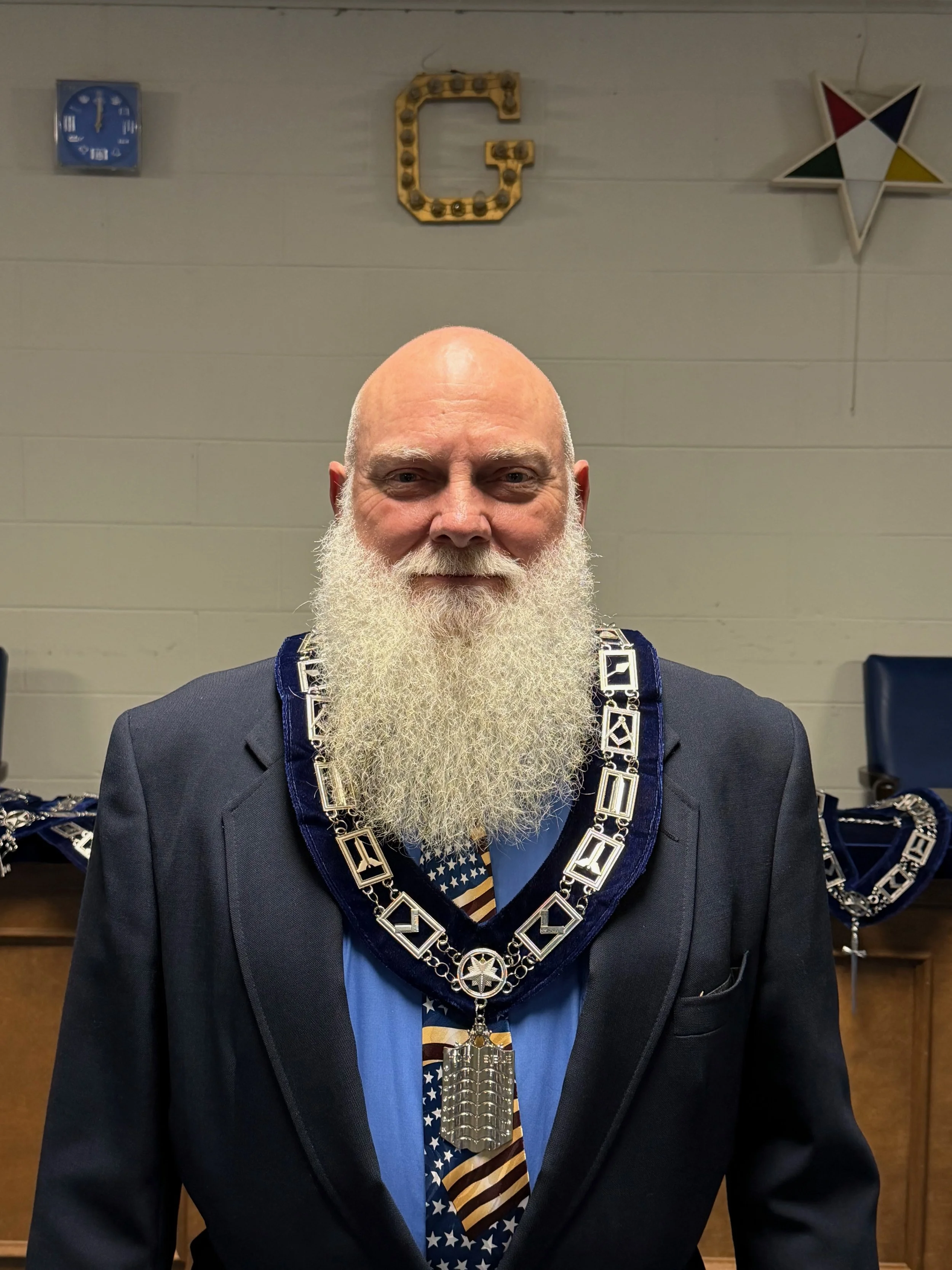 A man with a long white beard wearing a dark suit and a chain of office around his neck. Behind him are decorations including a large letter G made of lights, a star-shaped decoration with colored sections, and a clock on the wall.