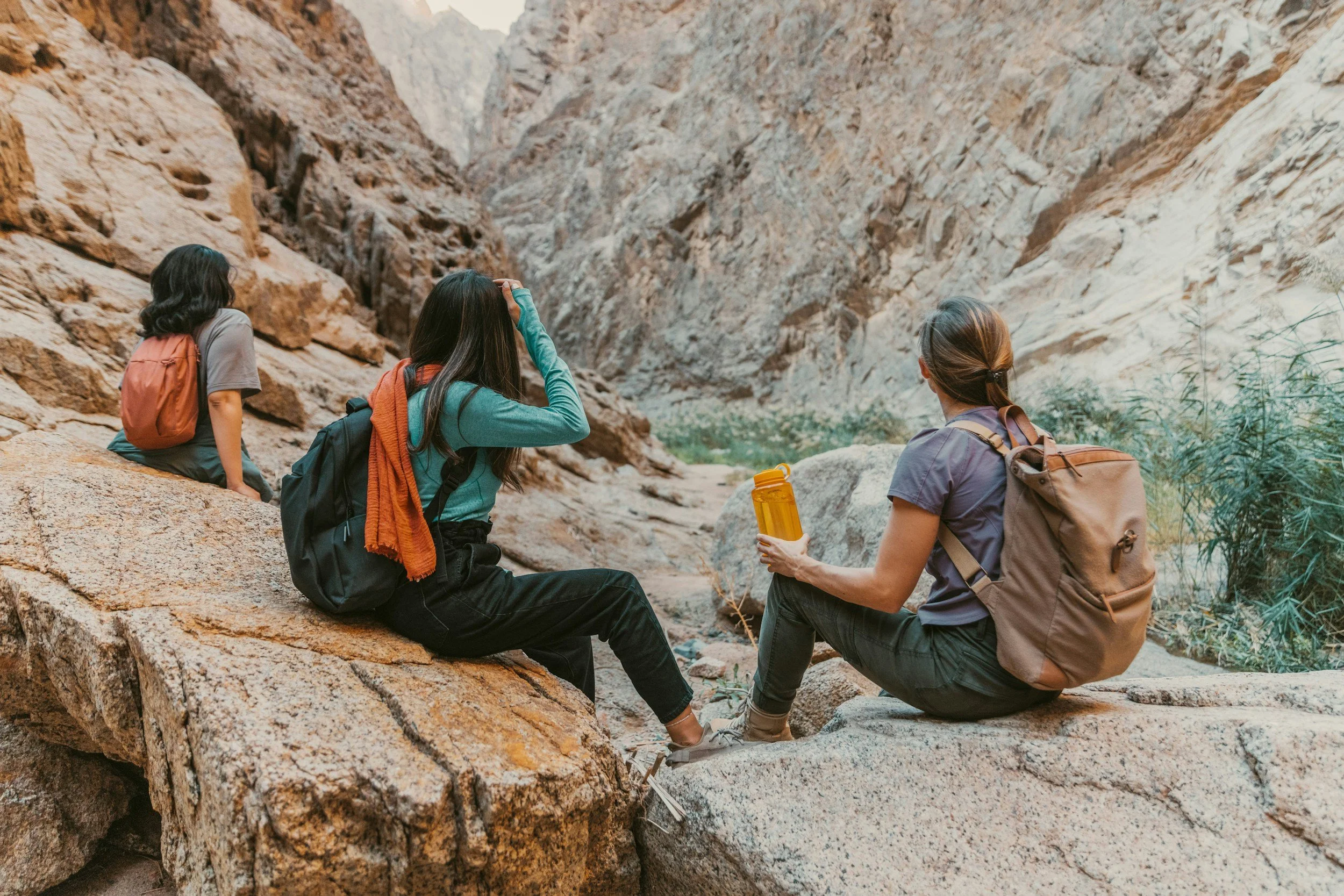 Three women with backpacks sitting on rocks in a canyon, resting during a hike.
