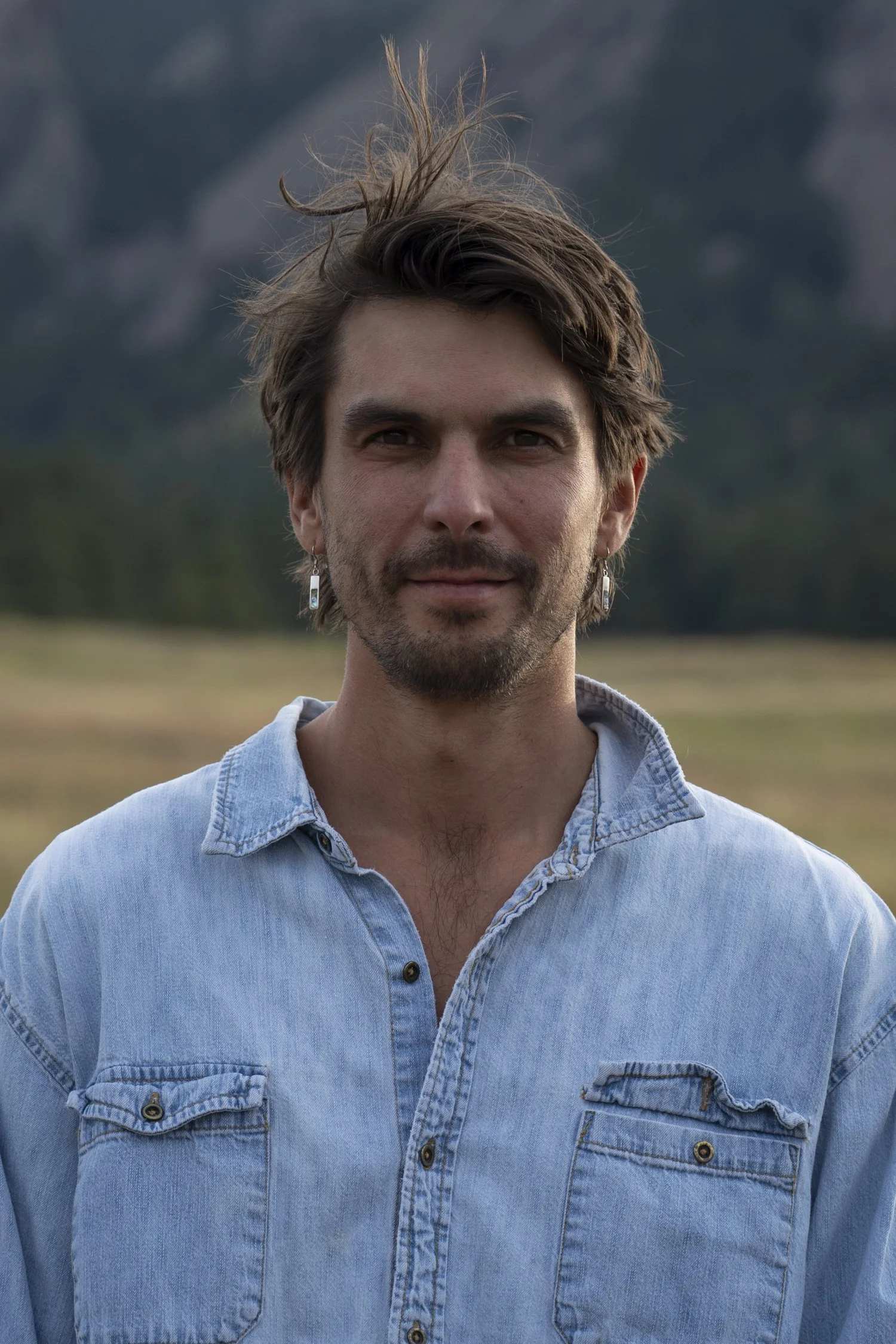 A man with brown hair and a beard, wearing a light blue denim shirt, standing outdoors in a natural landscape with mountains in the background.