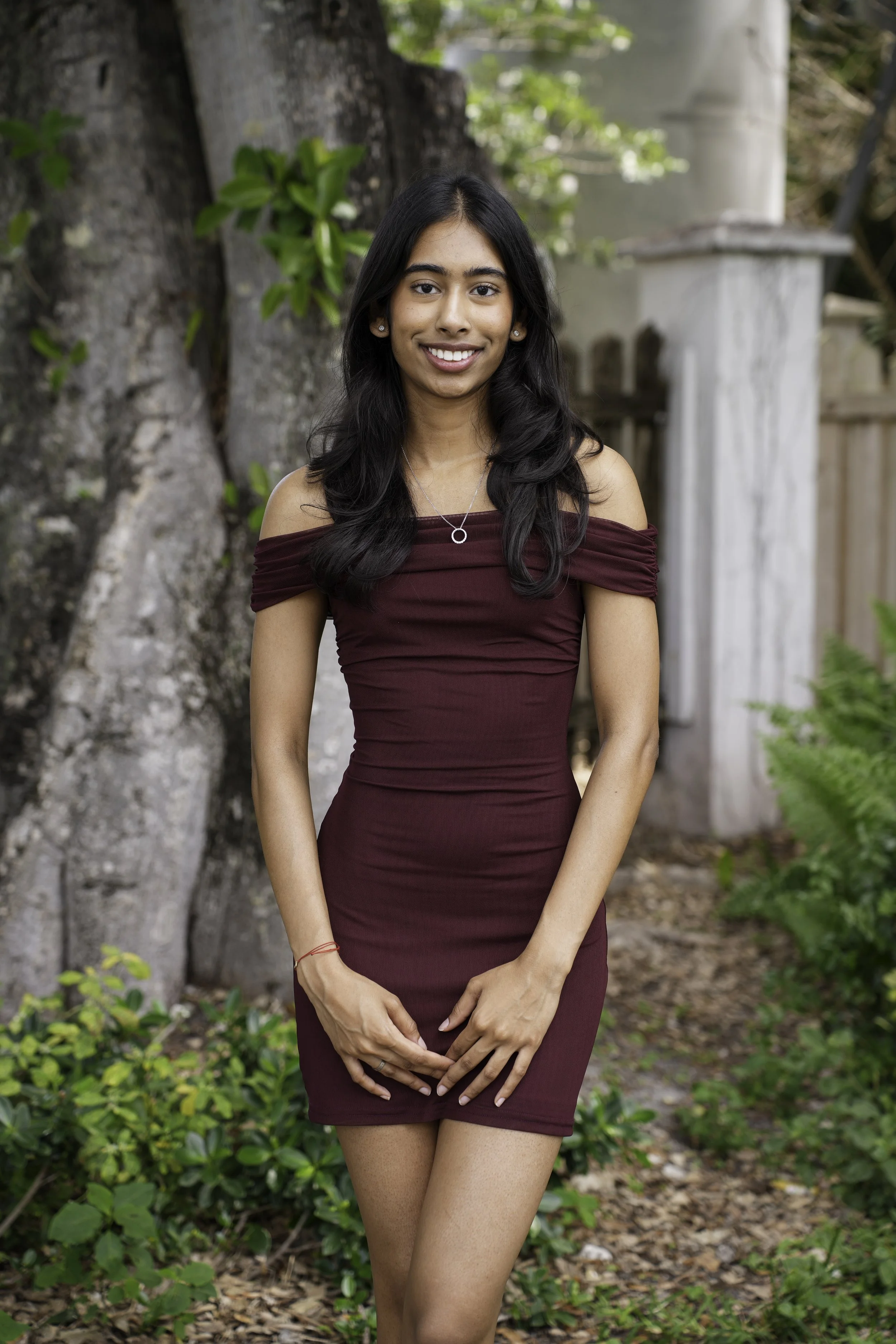 A young woman with long black hair, wearing a maroon off-shoulder dress, standing outdoors in front of a large tree and greenery.