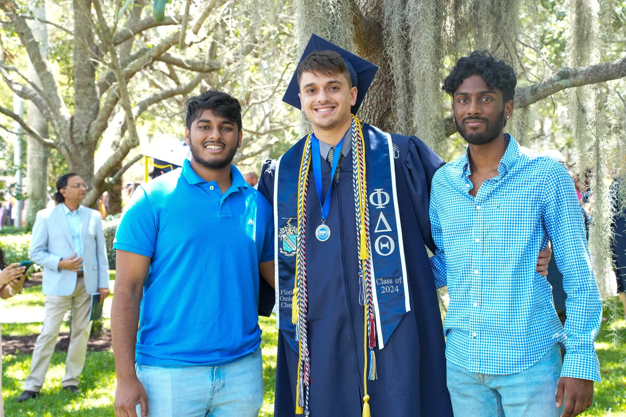 Graduation celebration with a man in a blue cap and gown with honor cords, and a woman in a light-colored dress, smiling outdoors.
