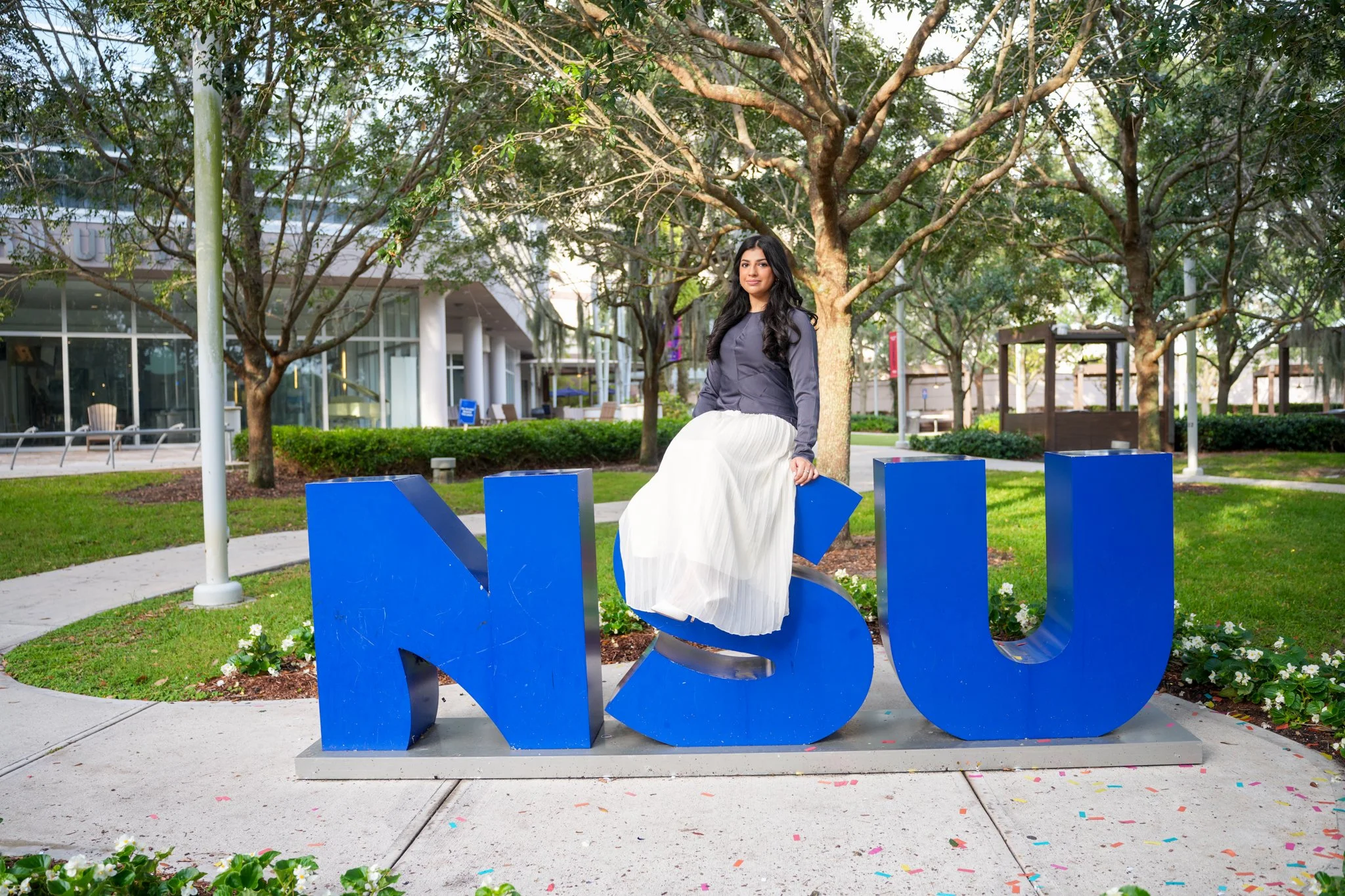 A woman sitting on a blue 'MSU' sign in a park with trees, grass, and a modern building in the background.